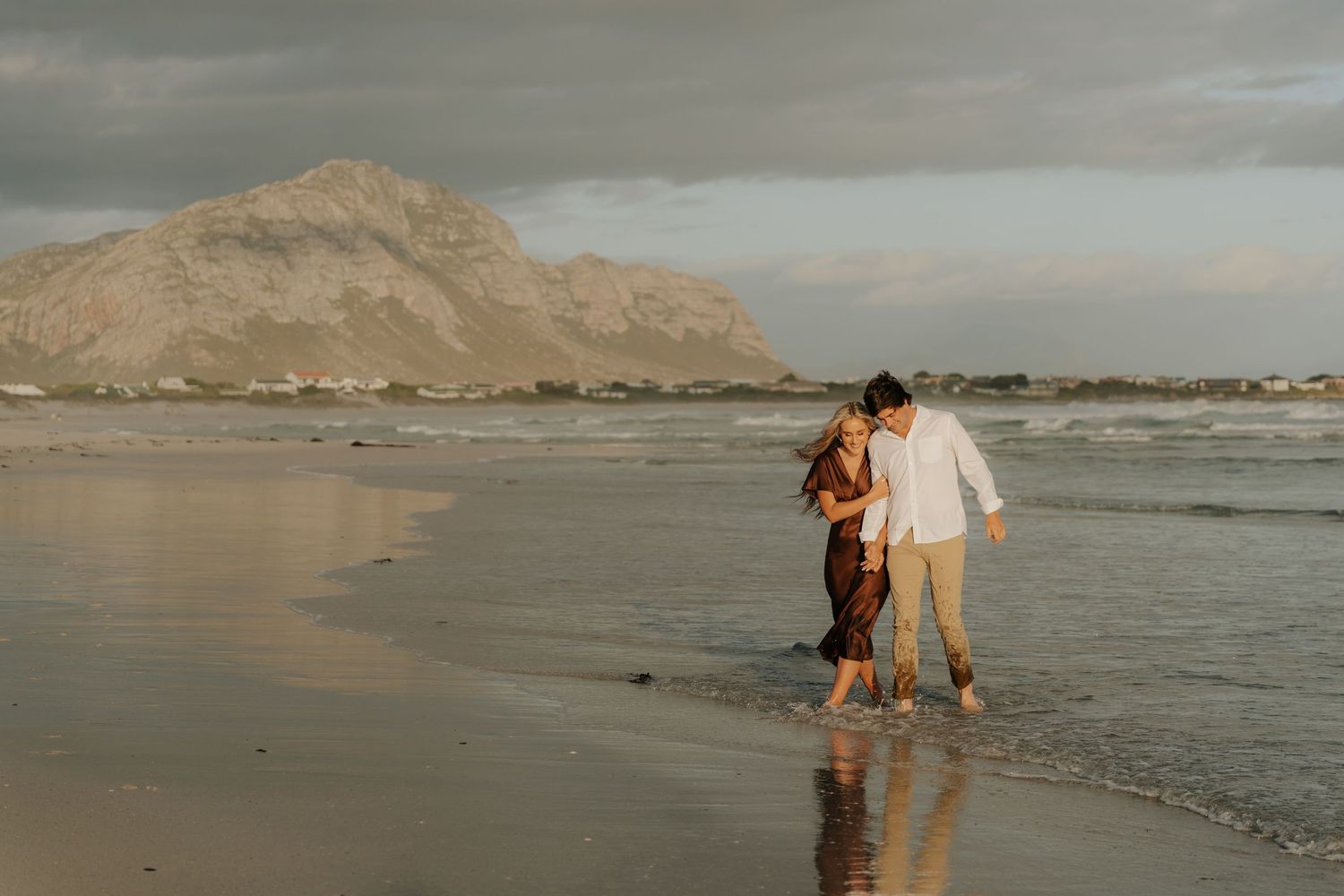 Couple walking barefoot along shoreline at golden hour with dramatic Cape Town mountain backdrop behind them.