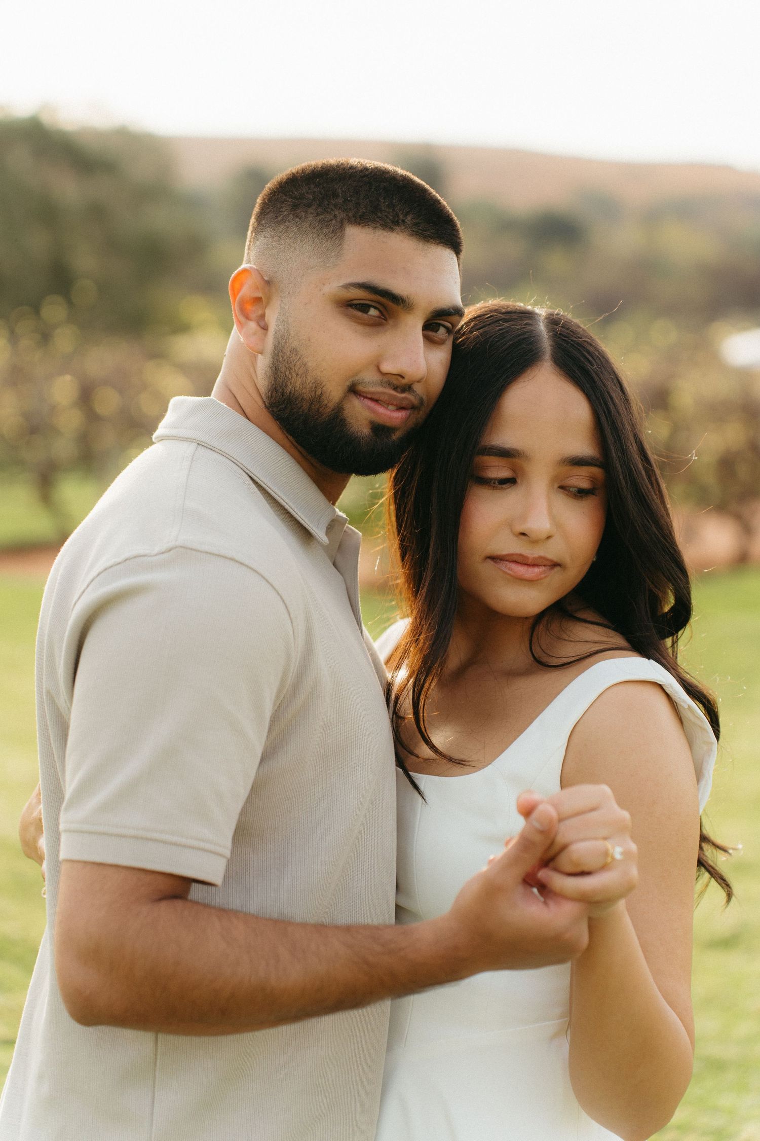 Man embraces woman from behind in sunlit field in Muldersdrift, both in neutral tones, holding hands tenderly.