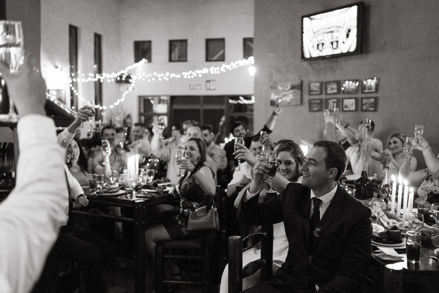 Black and white photo of wedding reception guests raising glasses for a toast in a decorated venue.