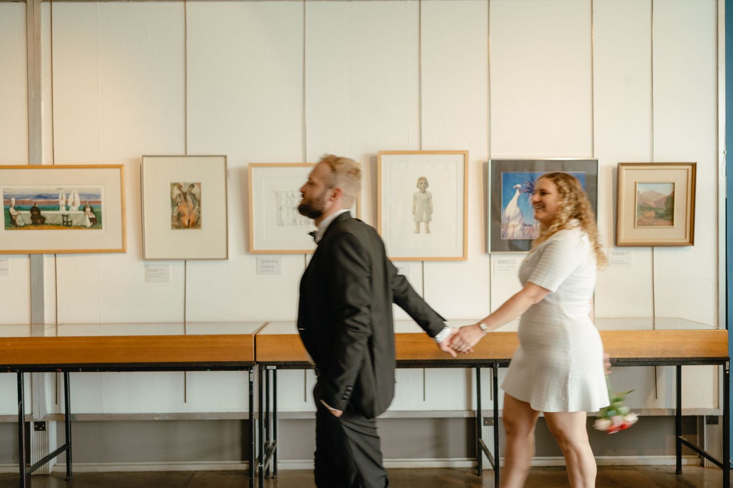 Couple in formal wear dances playfully among framed artwork in gallery space.