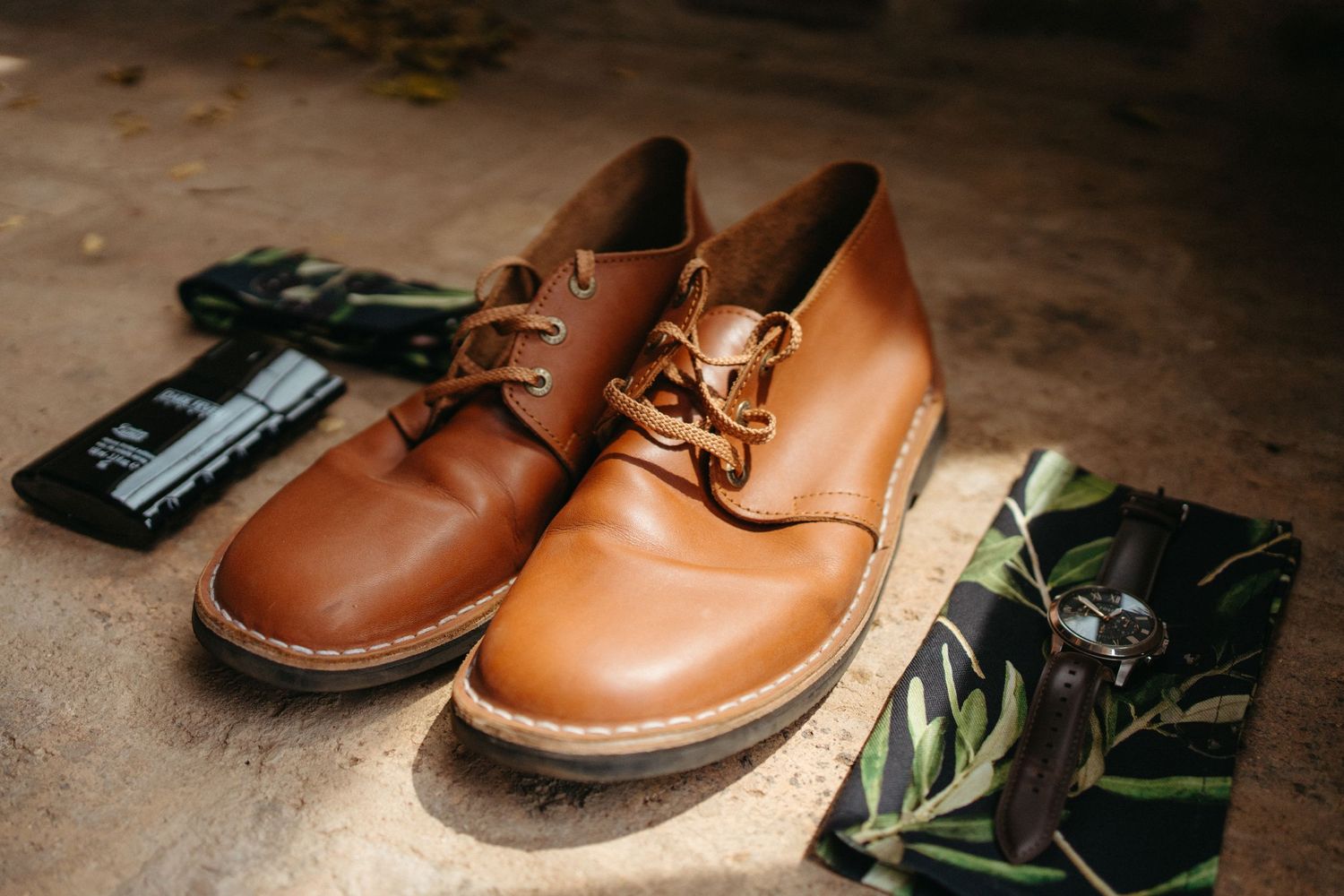 Brown leather dress shoes with botanical greenery and accessories arranged for wedding detail shot.