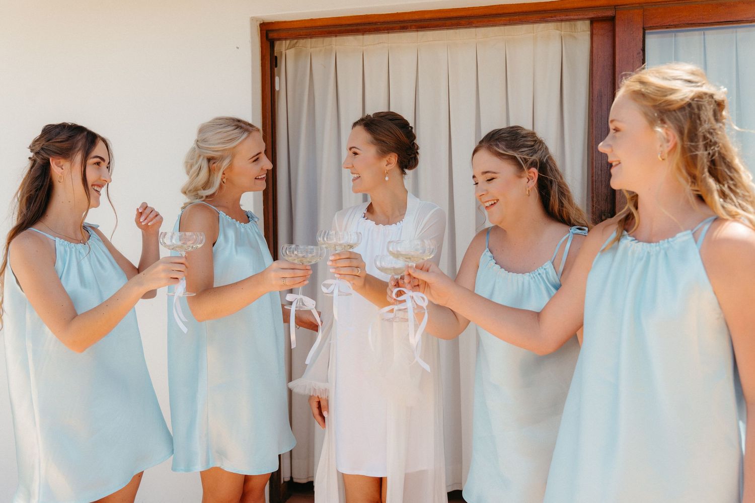 A group of women in light blue and white dresses stand in a row laughing and enjoying a celebratory moment together.
