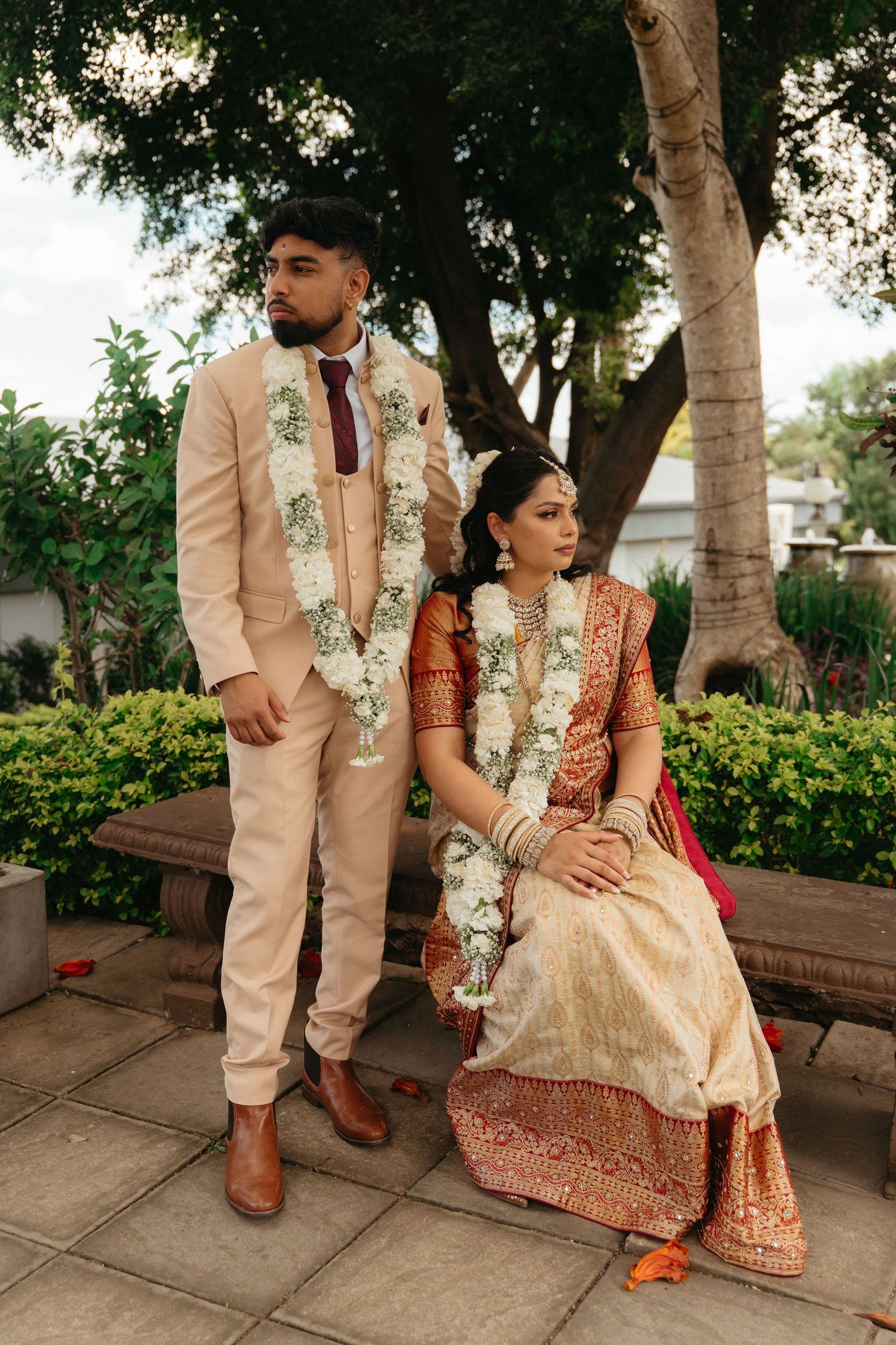 A couple in traditional South Asian wedding attire pose outdoors, wearing floral garlands and elegant formal clothing.