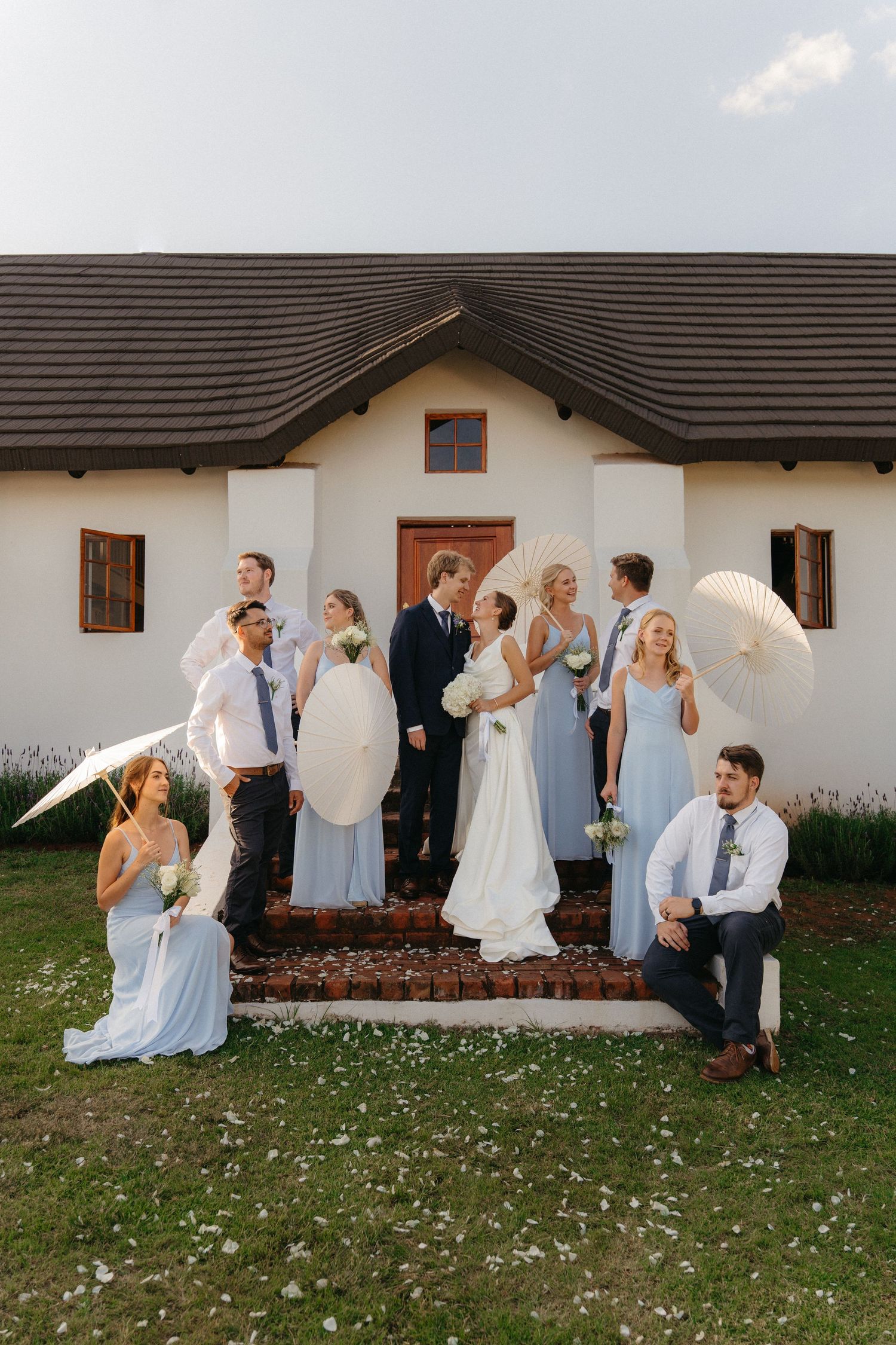 Wedding party in light blue and white attire poses playfully in sequence against white building exterior.