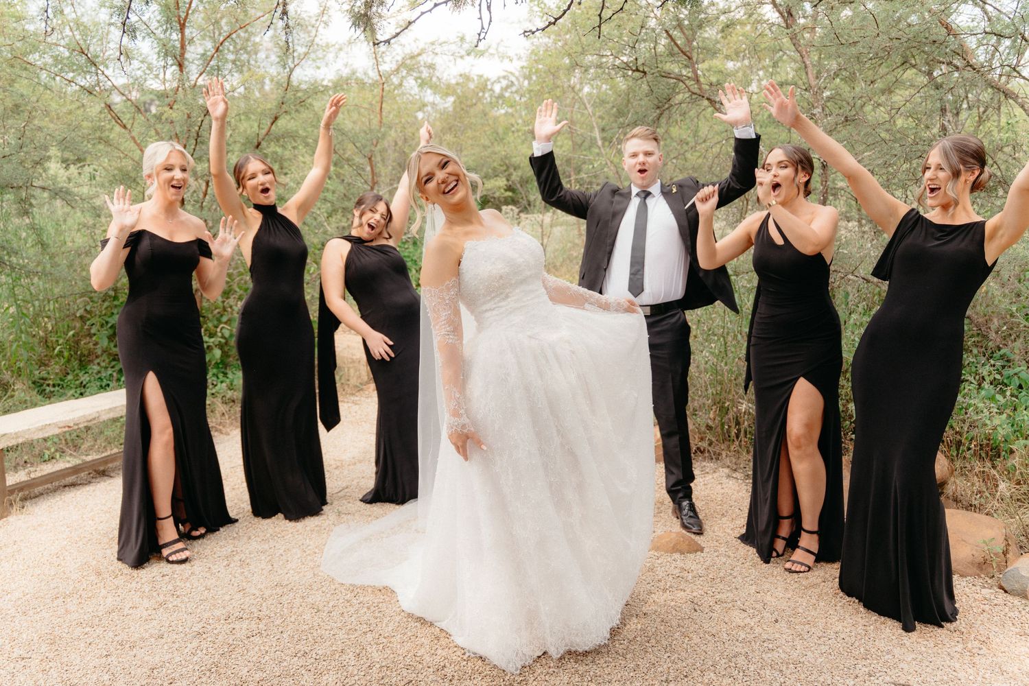 Wedding party in black dresses and white gown pose playfully together outdoors in a natural setting.