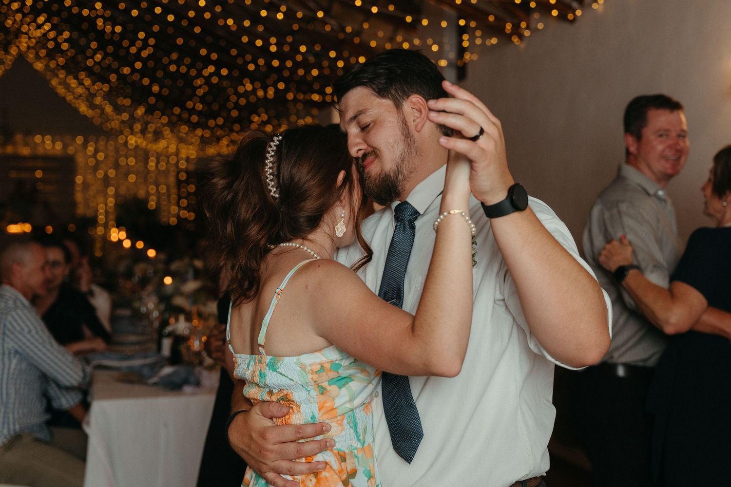 Couples dancing together at a wedding reception under warm glowing string lights and decorations.
