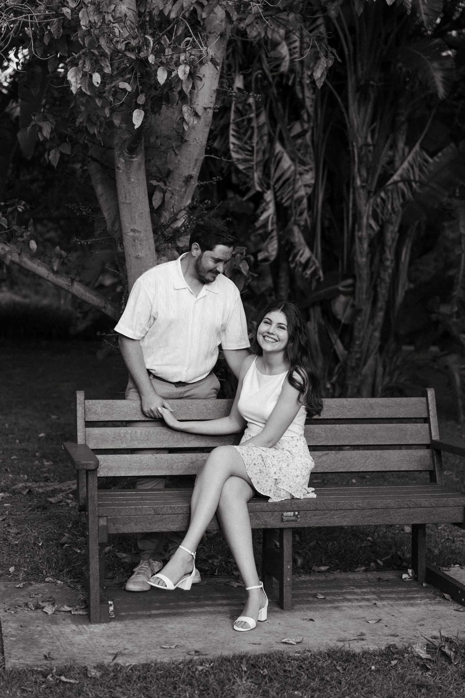 Black and white photo of a smiling couple on a wooden park bench surrounded by lush trees.