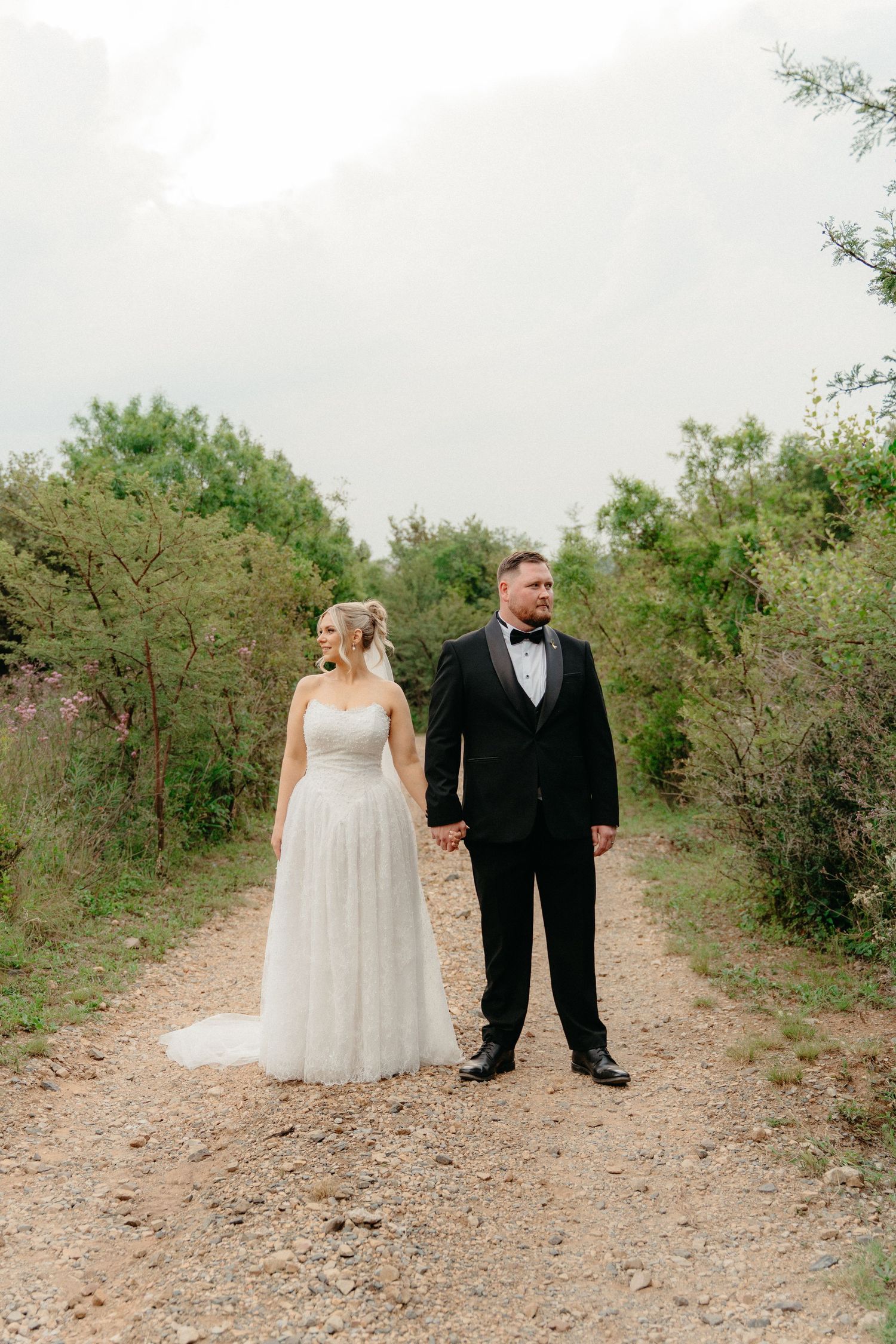 A couple in formal wedding attire stands together on a rustic dirt path surrounded by green foliage.