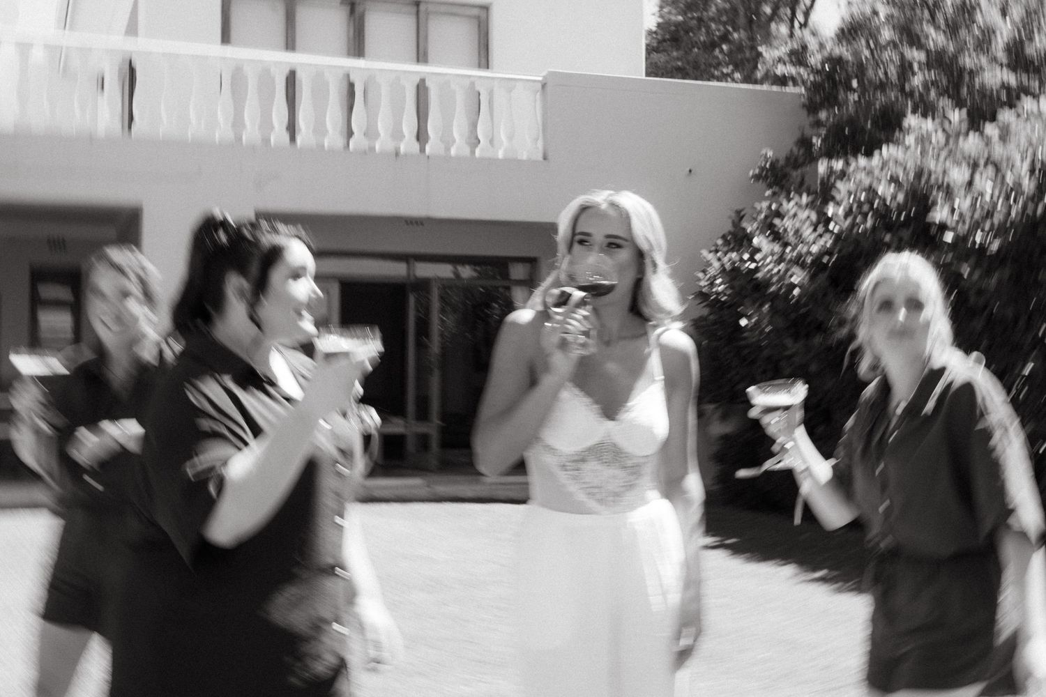 Black and white photo of a bride in a white dress laughing with champagne glasses among friends on a sunny patio at Duvon Wine Estate.