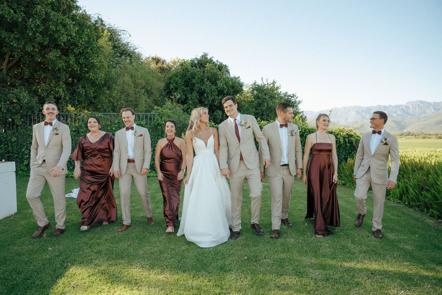 Bride in a white gown walks across a sunny lawn with groomsmen in tan suits and bridesmaids in brown dresses, mountains in the background.