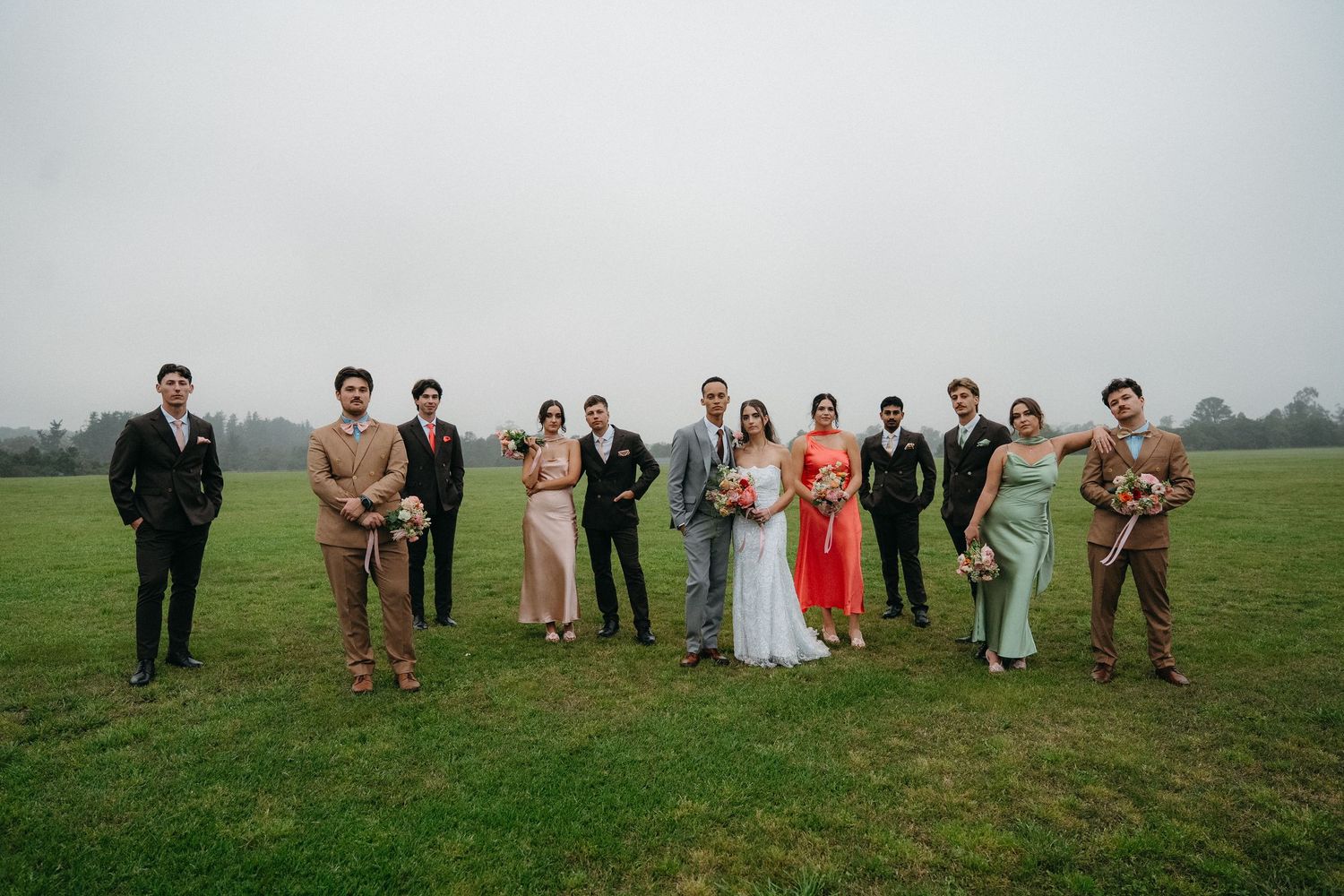 Wedding party poses on misty green Western Cape field, bride in red, groom in grey, bridesmaids and groomsmen in elegant attire.
