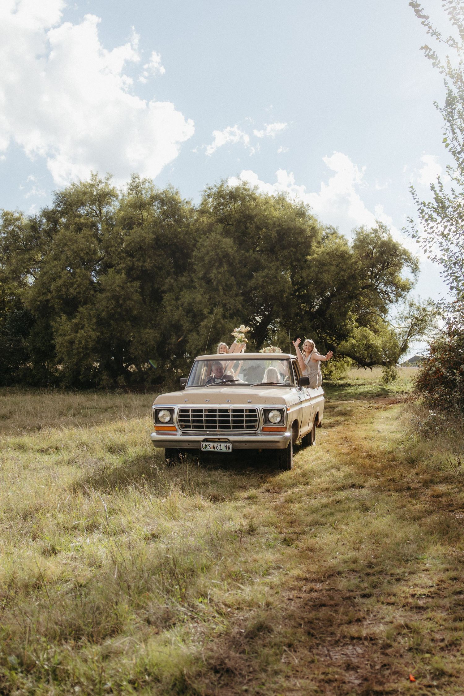 Friends joyfully riding in a vintage white pickup truck through a scenic rural field with lush green trees under a cloudy sky.