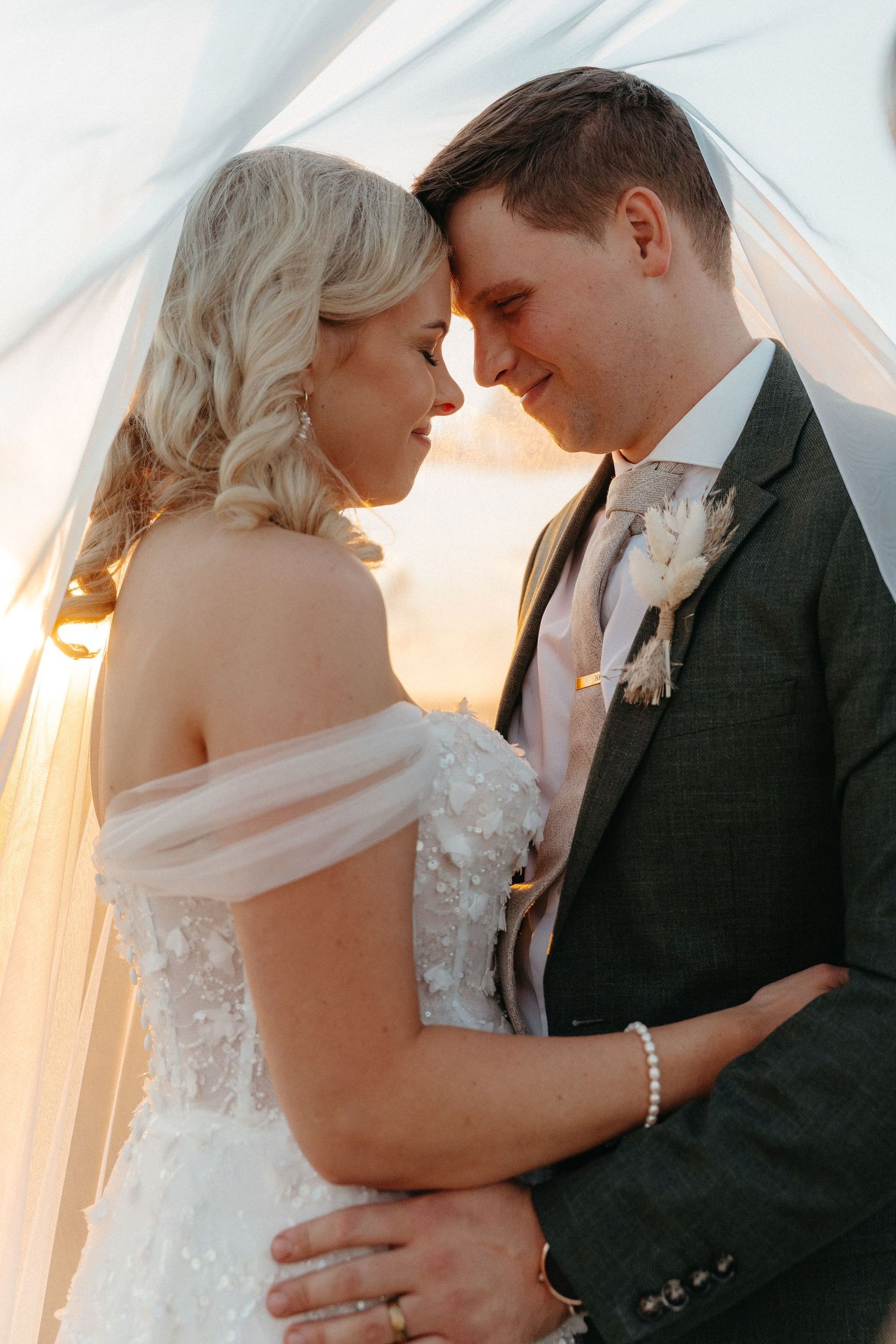 Bride and groom share an intimate moment under a veil at golden hour, bride in off-shoulder gown, groom in gray suit.