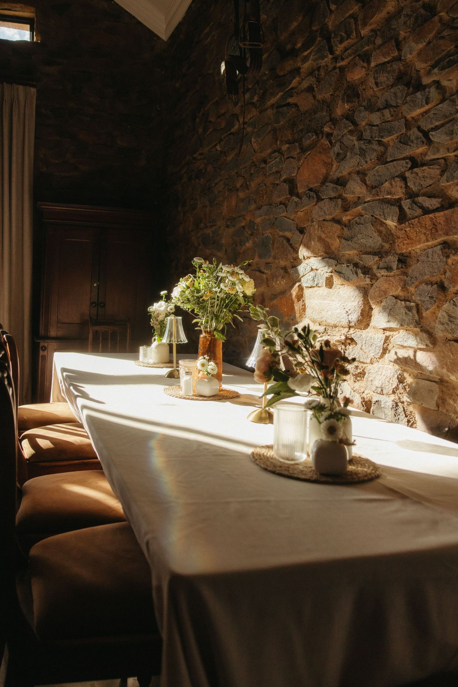 Sunlit dining table with white linen and floral centerpieces against a rustic stone wall.