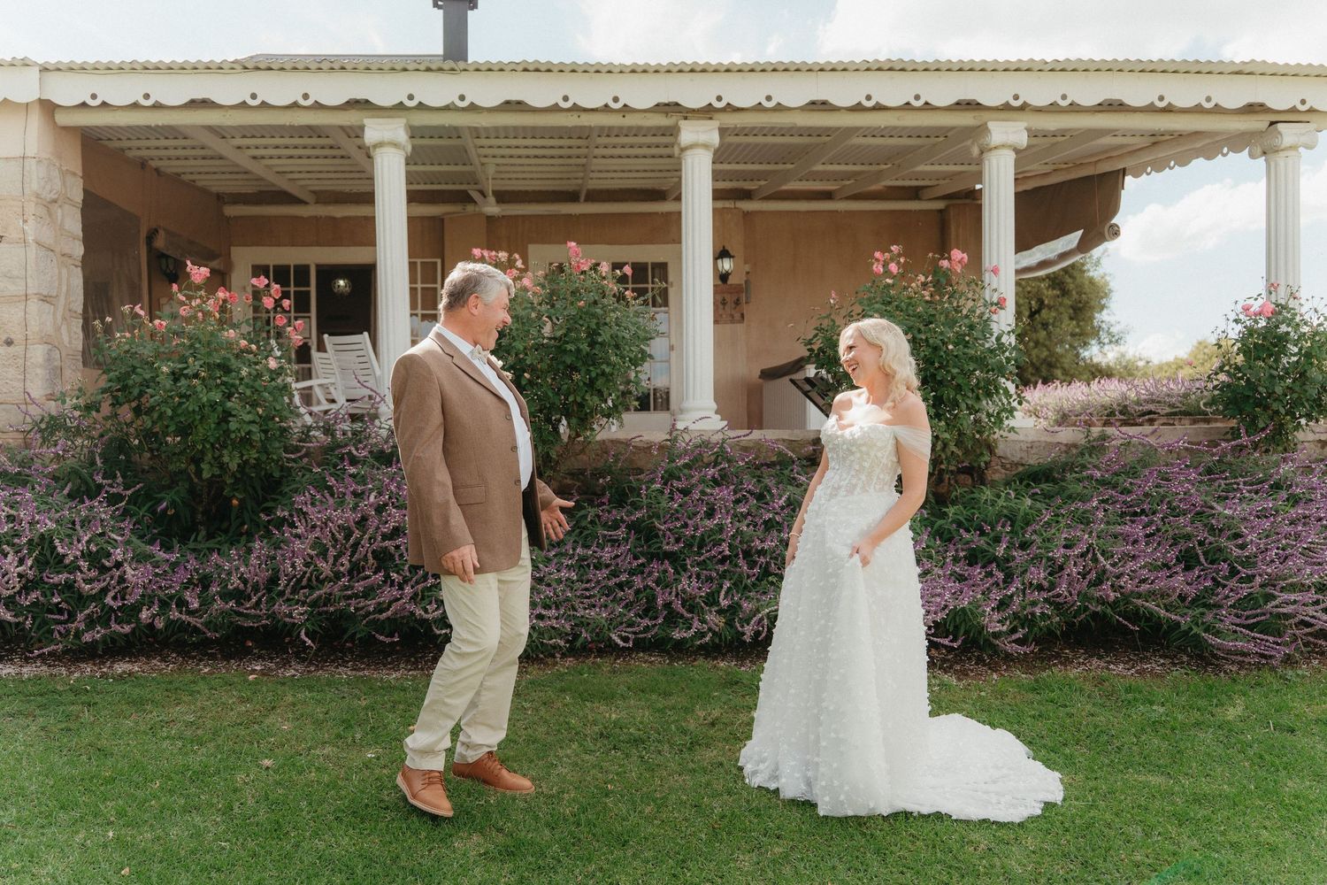 Bride in white lace gown and groom in tan suit share first look moment on green lawn outside rustic venue.