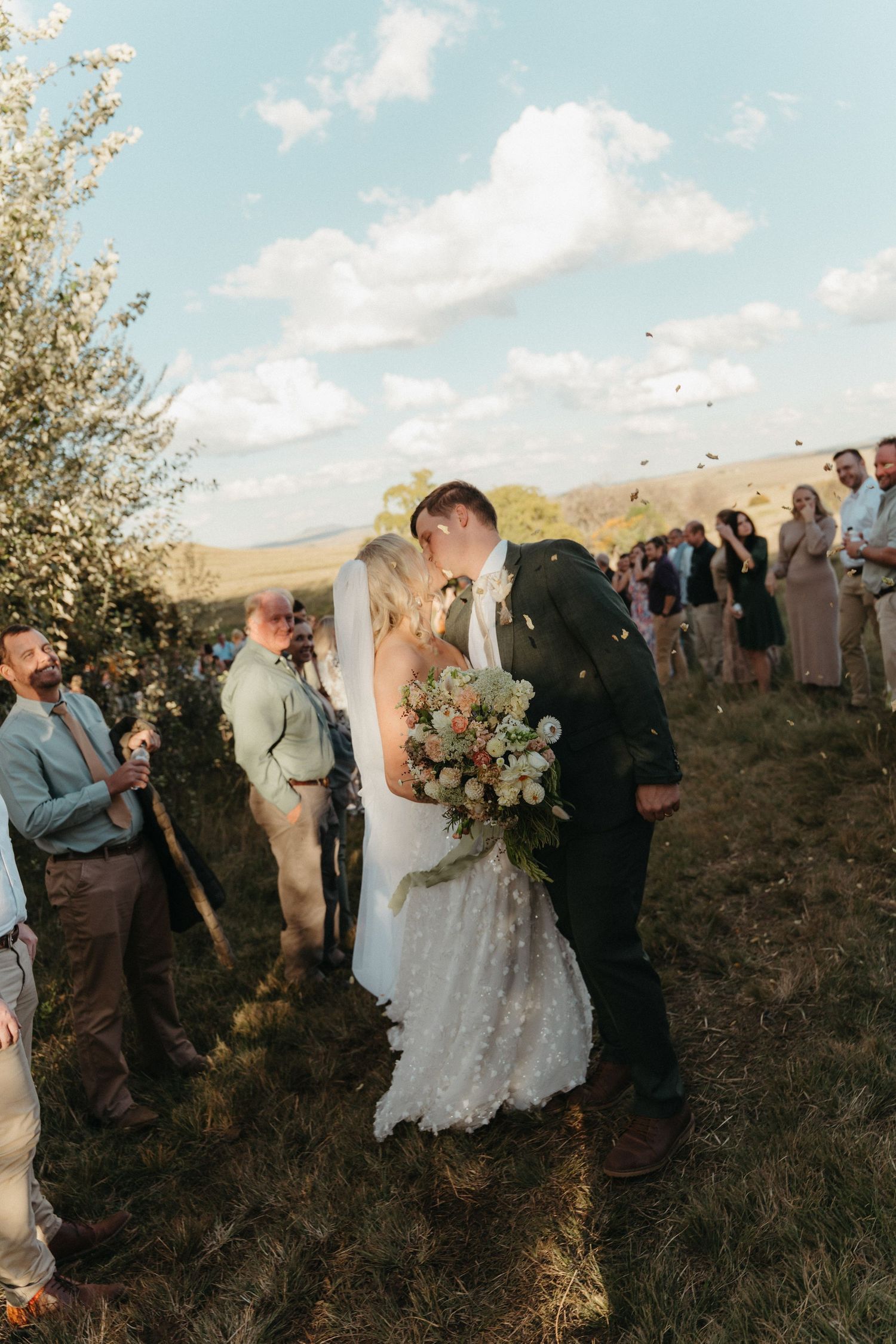Bride and groom share a kiss at outdoor wedding ceremony while guests celebrate with confetti on a sunny day.