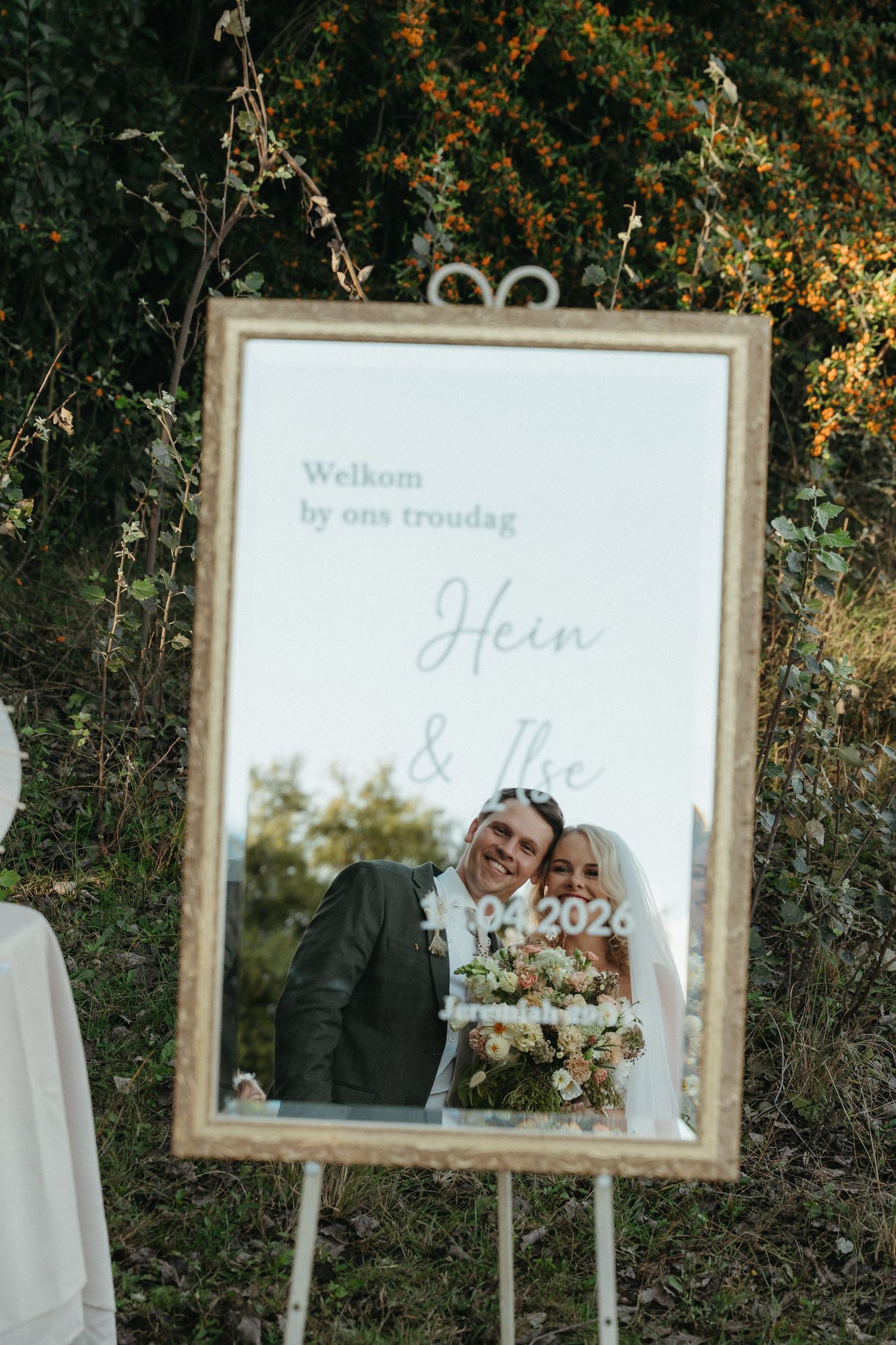 A wedding welcome mirror sign reading Hein and Ilse with couple reflection, displayed on easel outdoors.