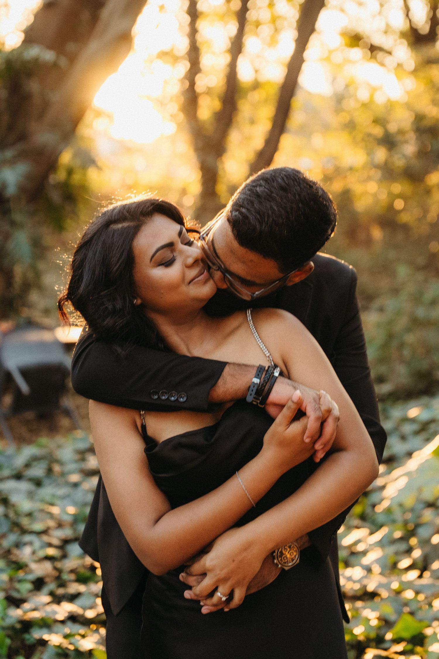 Couple in black outfits embracing in golden sunset light among trees, man kissing woman's cheek romantically.