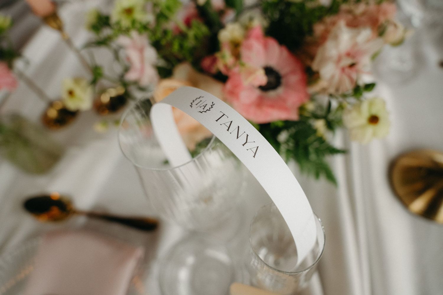 Elegant wedding place card reading TANYA on a glass vase surrounded by pink floral arrangements and gold cutlery.