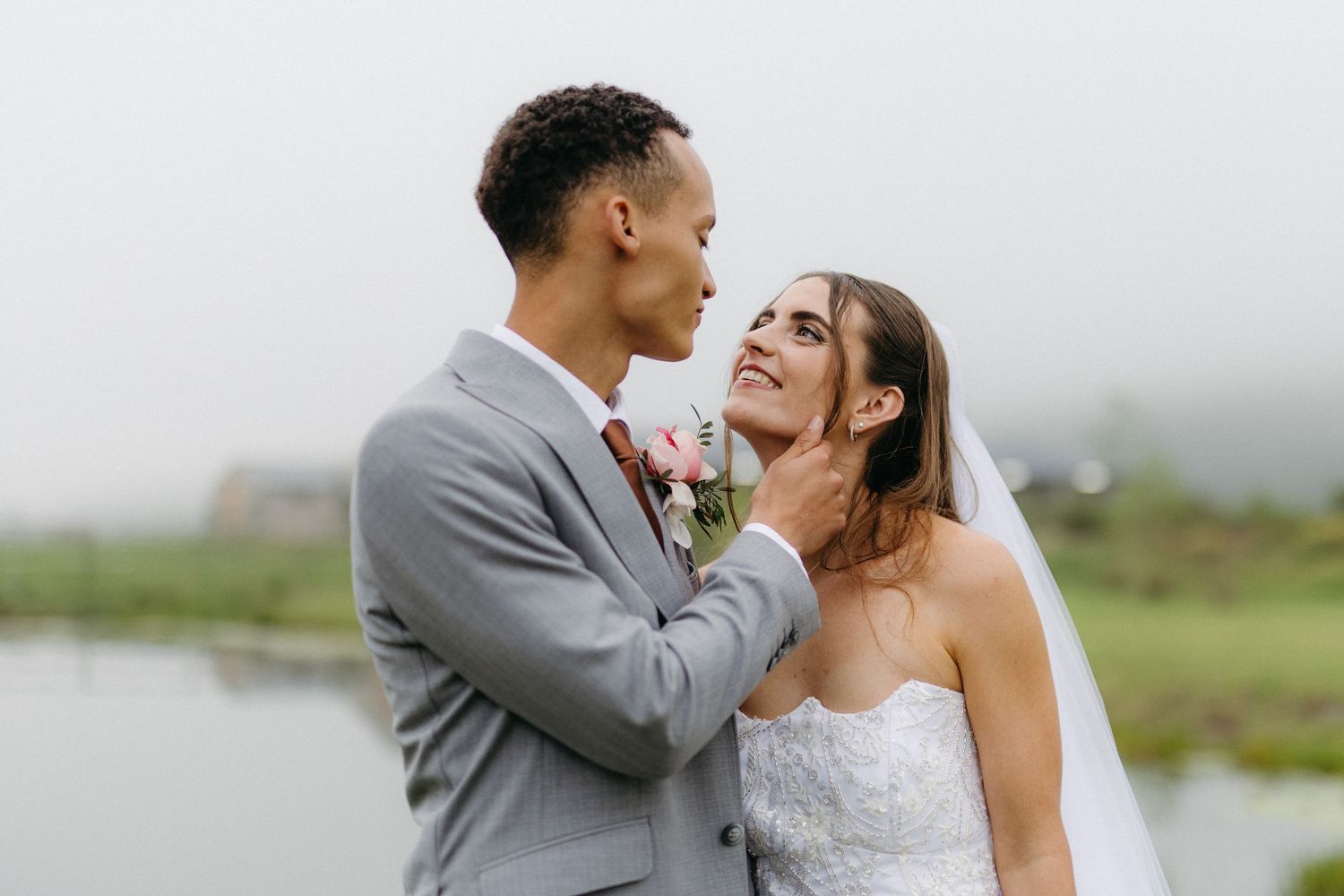 A groom in a gray suit tenderly holds his bride's face as they share a romantic moment outdoors on their wedding day.