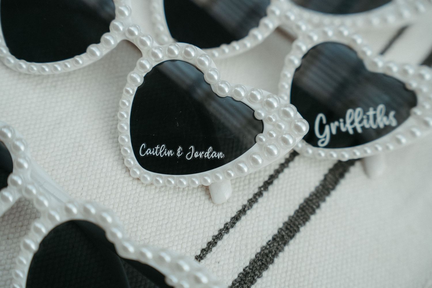 Heart-shaped pearl-rimmed sunglasses personalized with names Caitlin & Jordan and Griffiths for a wedding favor.