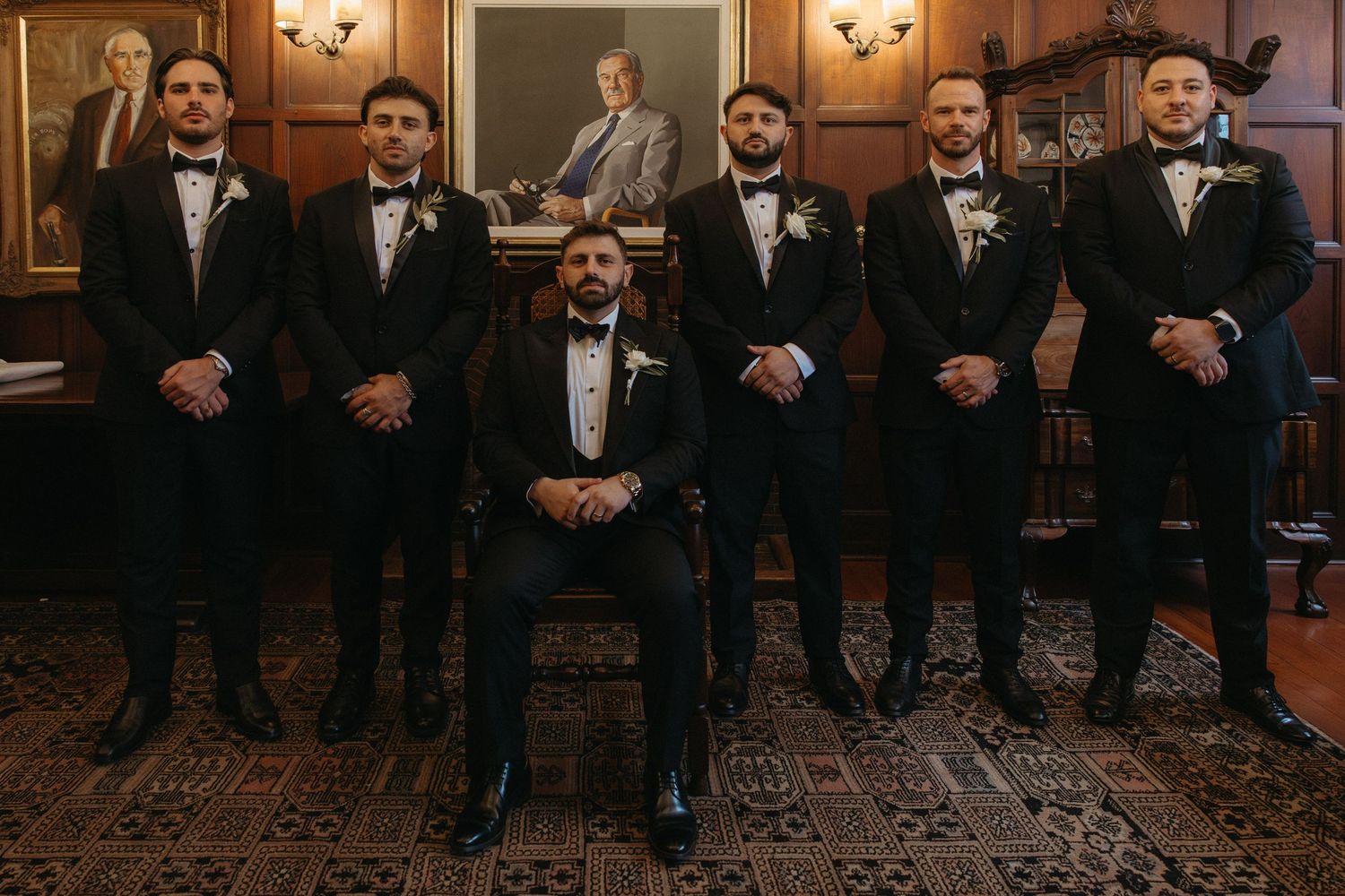 Groom seated with six groomsmen in black tuxedos and white boutonnieres posing in an elegant wood-paneled room.