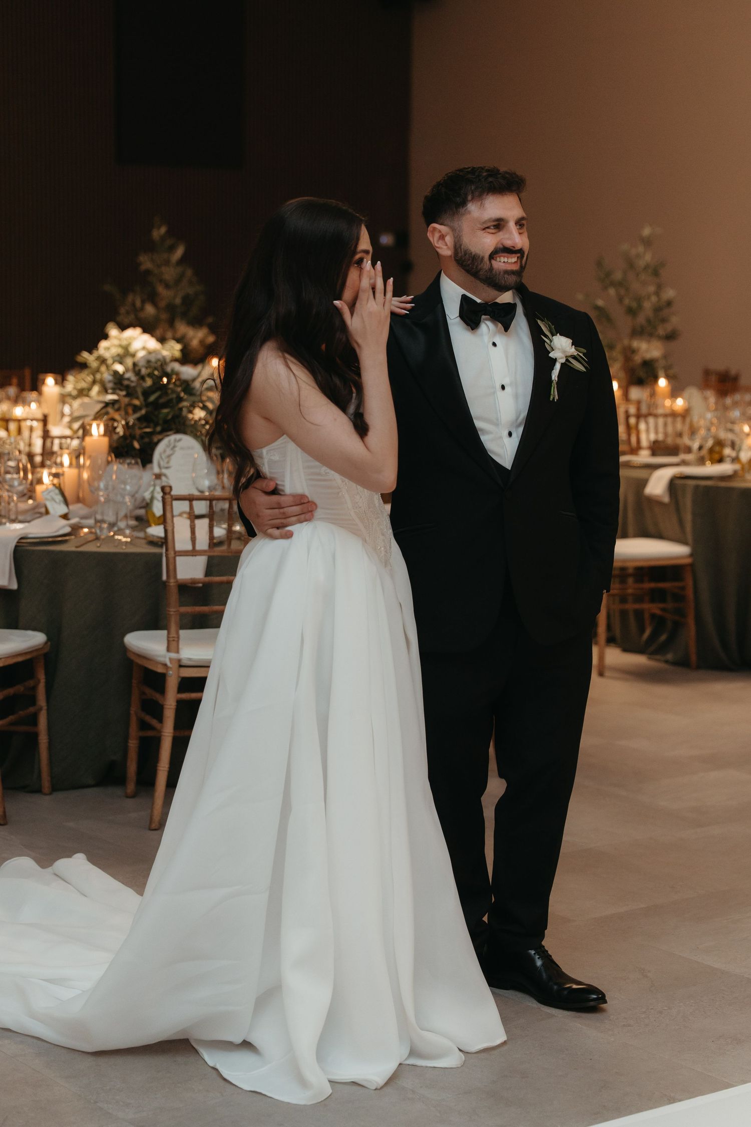 Bride in white gown wipes tears beside groom in black tuxedo at elegant candlelit wedding reception.