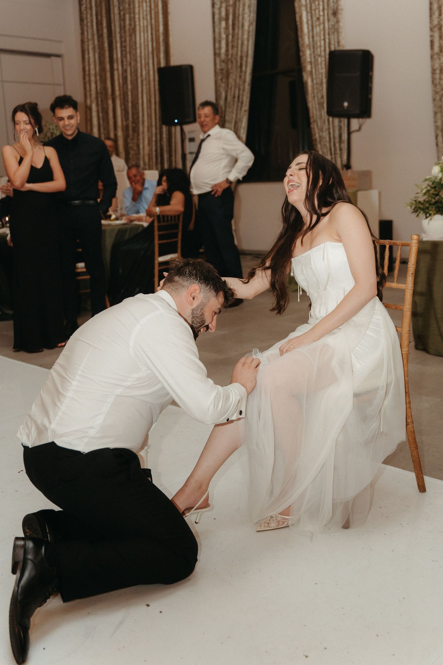 Groom kneels on dance floor to put shoes on laughing bride at elegant wedding reception.