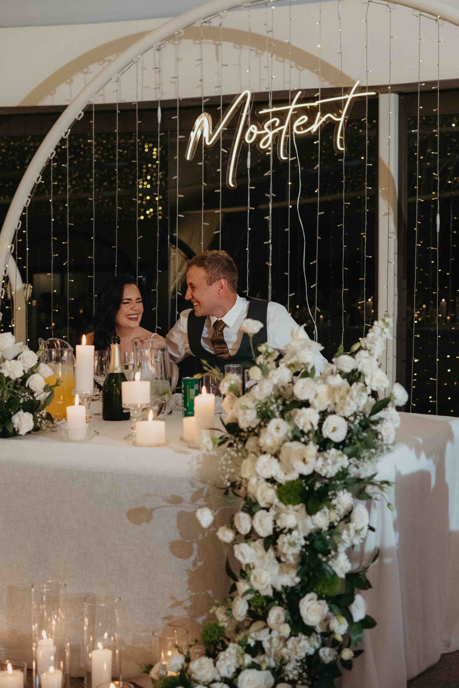 Couple smiling at wedding sweetheart table with white floral arrangements, candles, and Mostert neon sign backdrop.