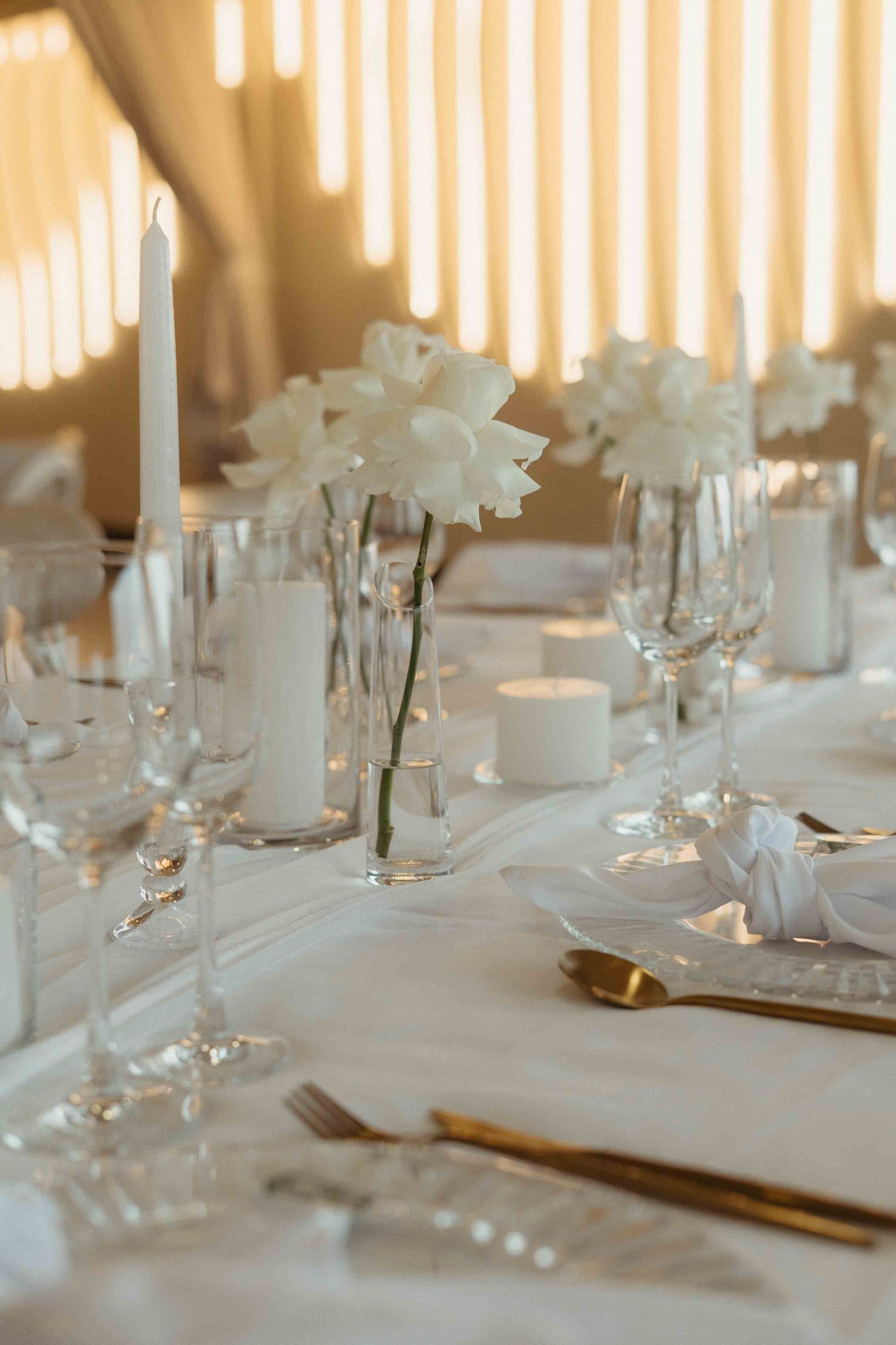 Elegant white wedding table setting with crystal glasses, gold cutlery, white flowers, and candles on a white tablecloth.