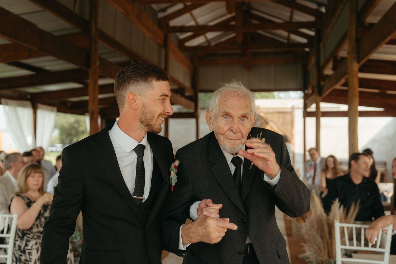 Two people share an emotional moment at an indoor venue with wooden beams and white chairs.