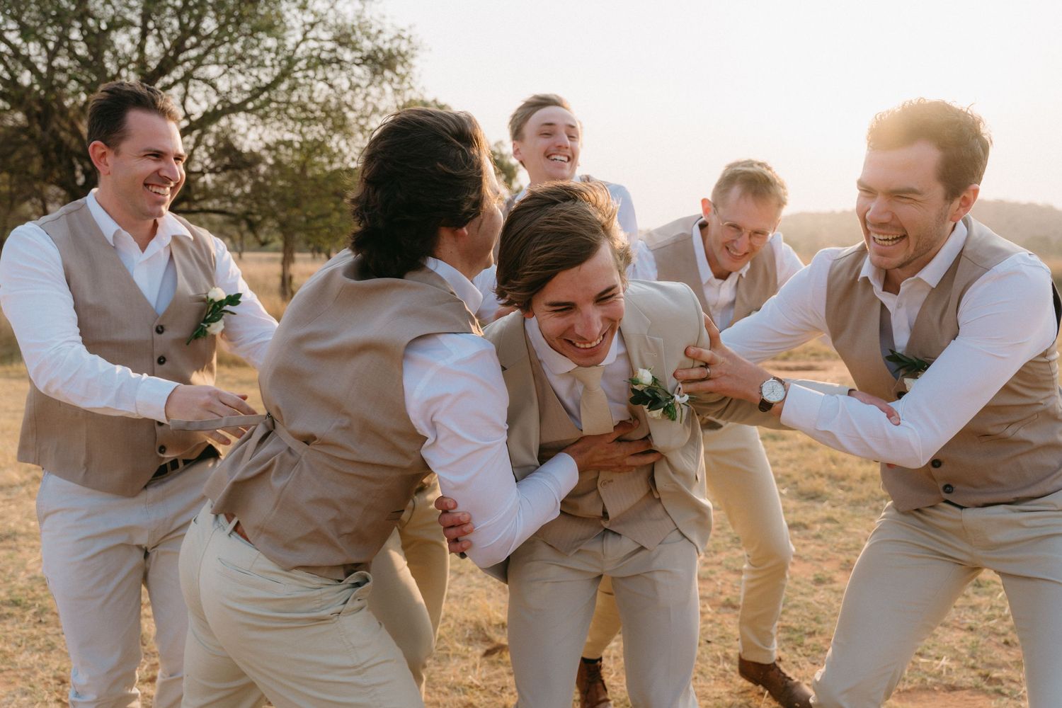 Groomsmen in tan vests and white shirts laugh together while posing for candid photos outdoors.