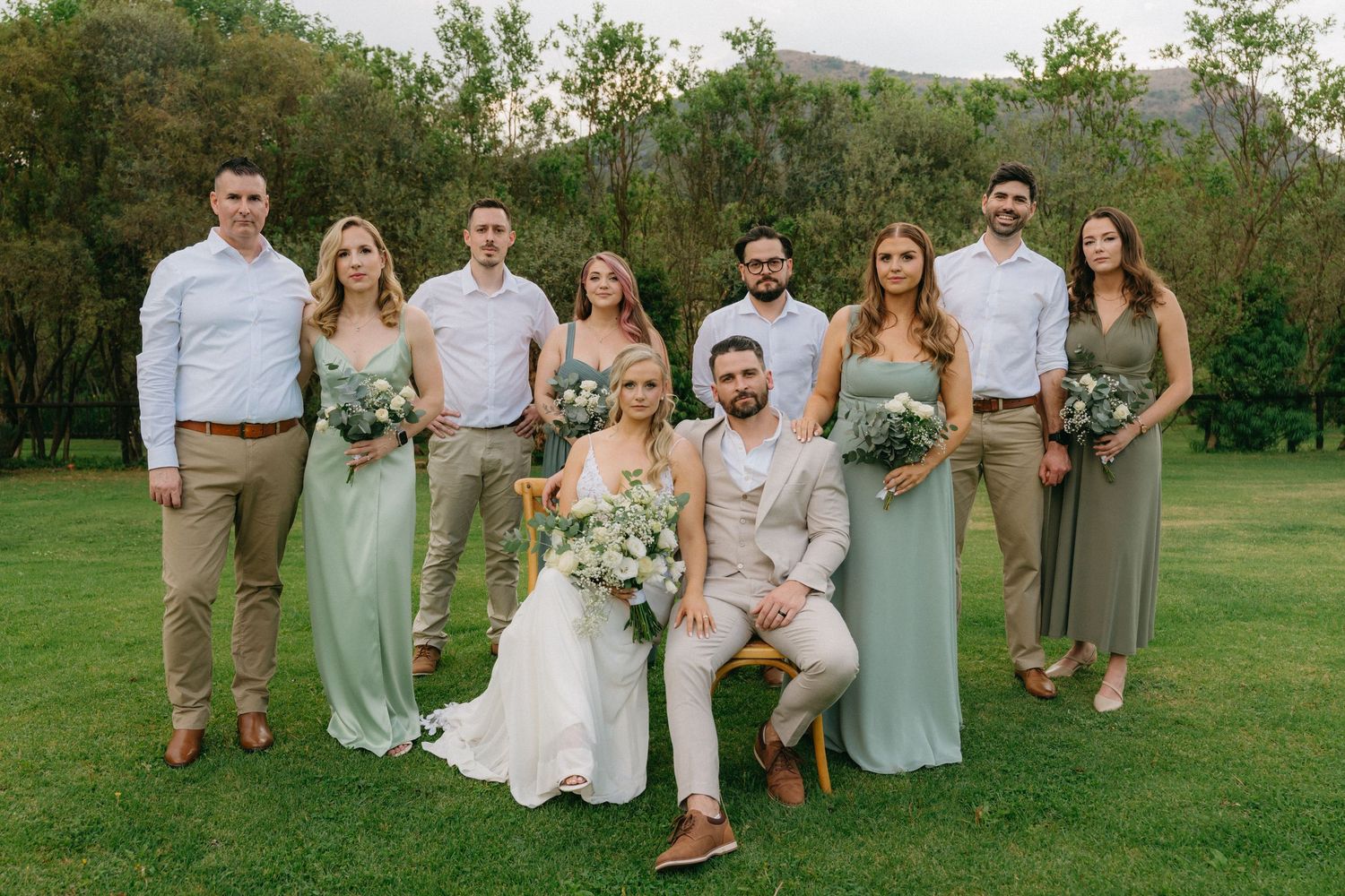 Wedding party in sage and neutral tones poses on green lawn.