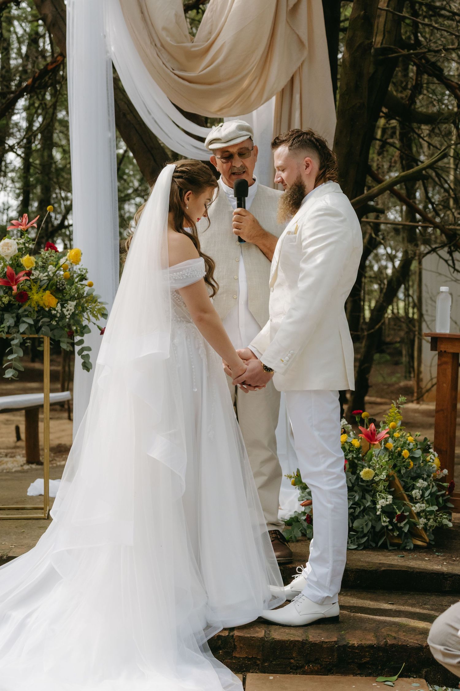 Wedding ceremony under draped fabric arch with colorful floral arrangements in an outdoor woodland setting.