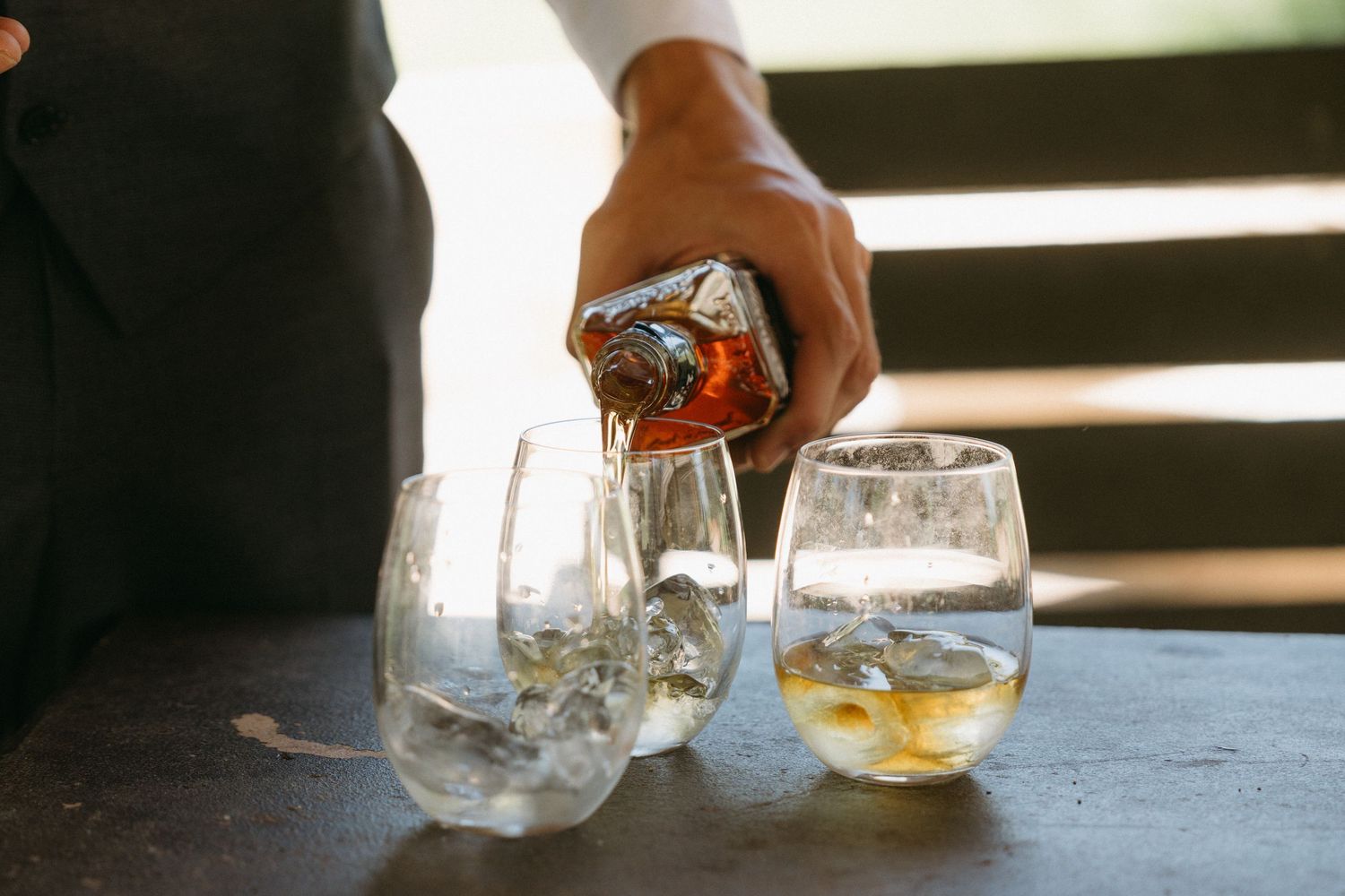Hand pouring a cocktail into glasses with ice at an outdoor bar setting.