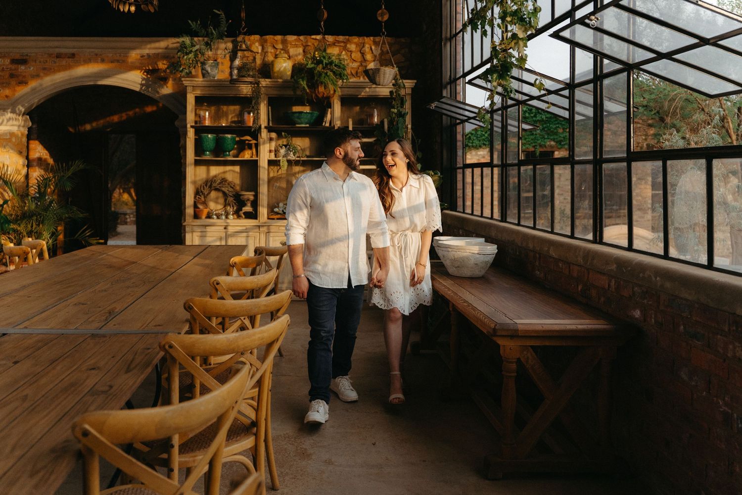 Couple in white outfits walks through a rustic greenhouse with wooden furniture and natural lighting.