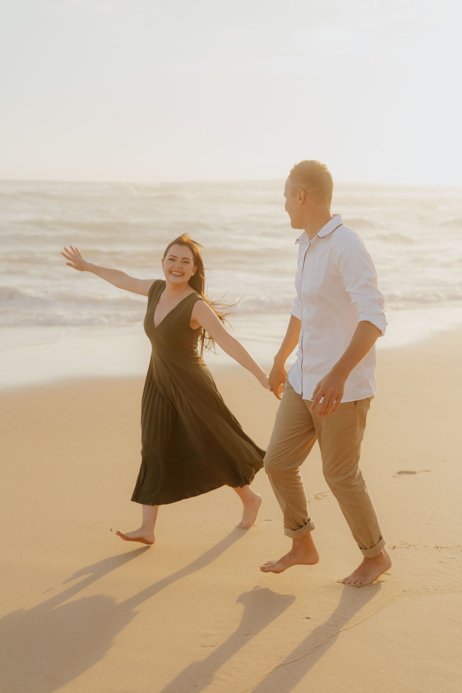 Couple dancing playfully on a sunlit Dana Bay Beach in Mossel Bay at sunset, casting long shadows in the sand.