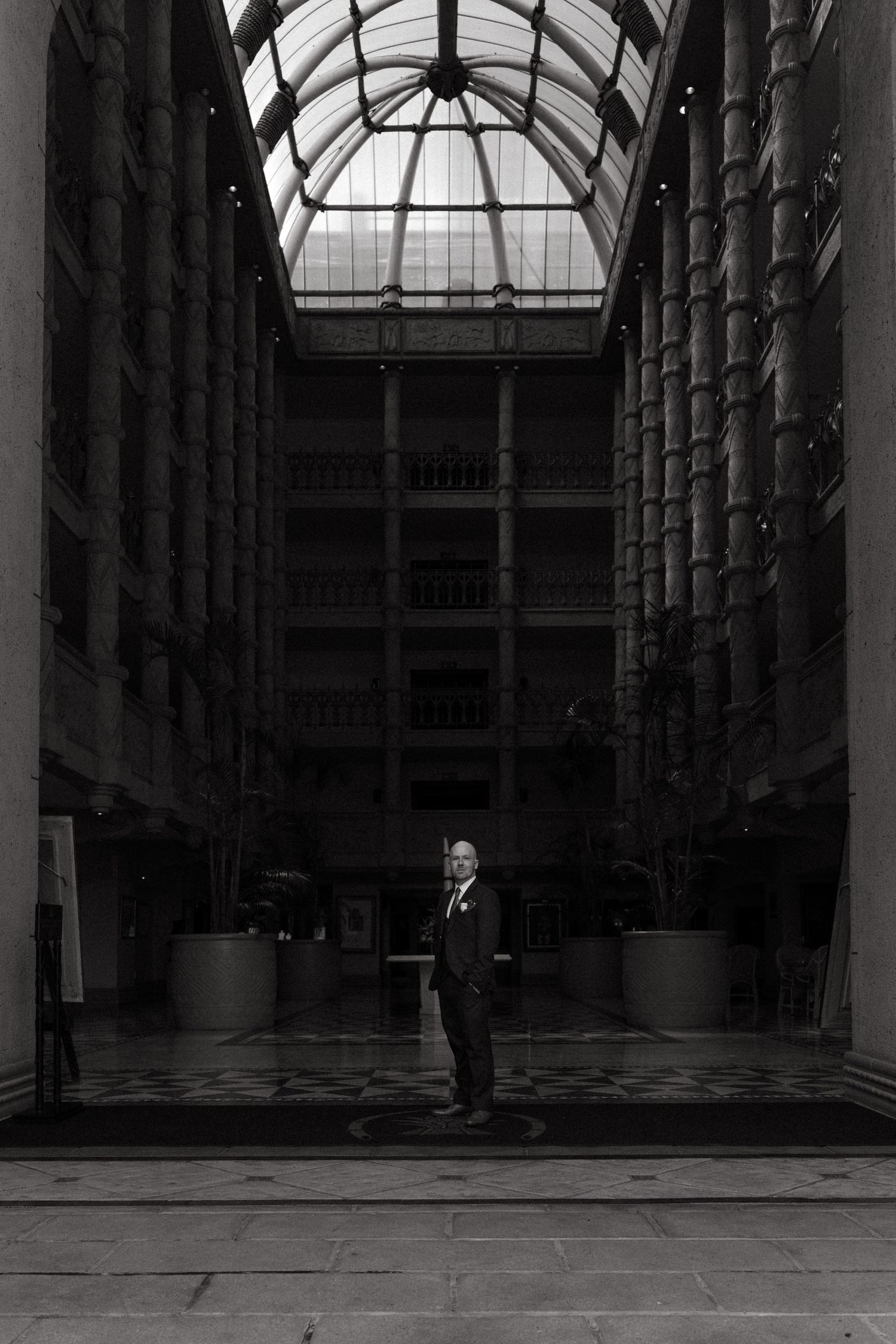A black and white architectural photo showing a person standing in a grand atrium with a glass ceiling and tall columns.