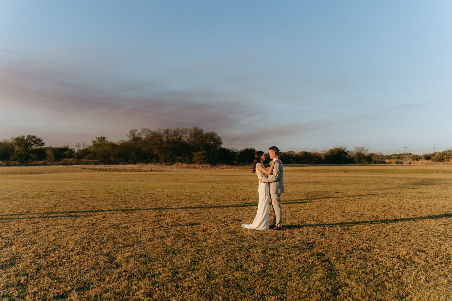 A couple in wedding attire stands together in a vast golden field at sunset.