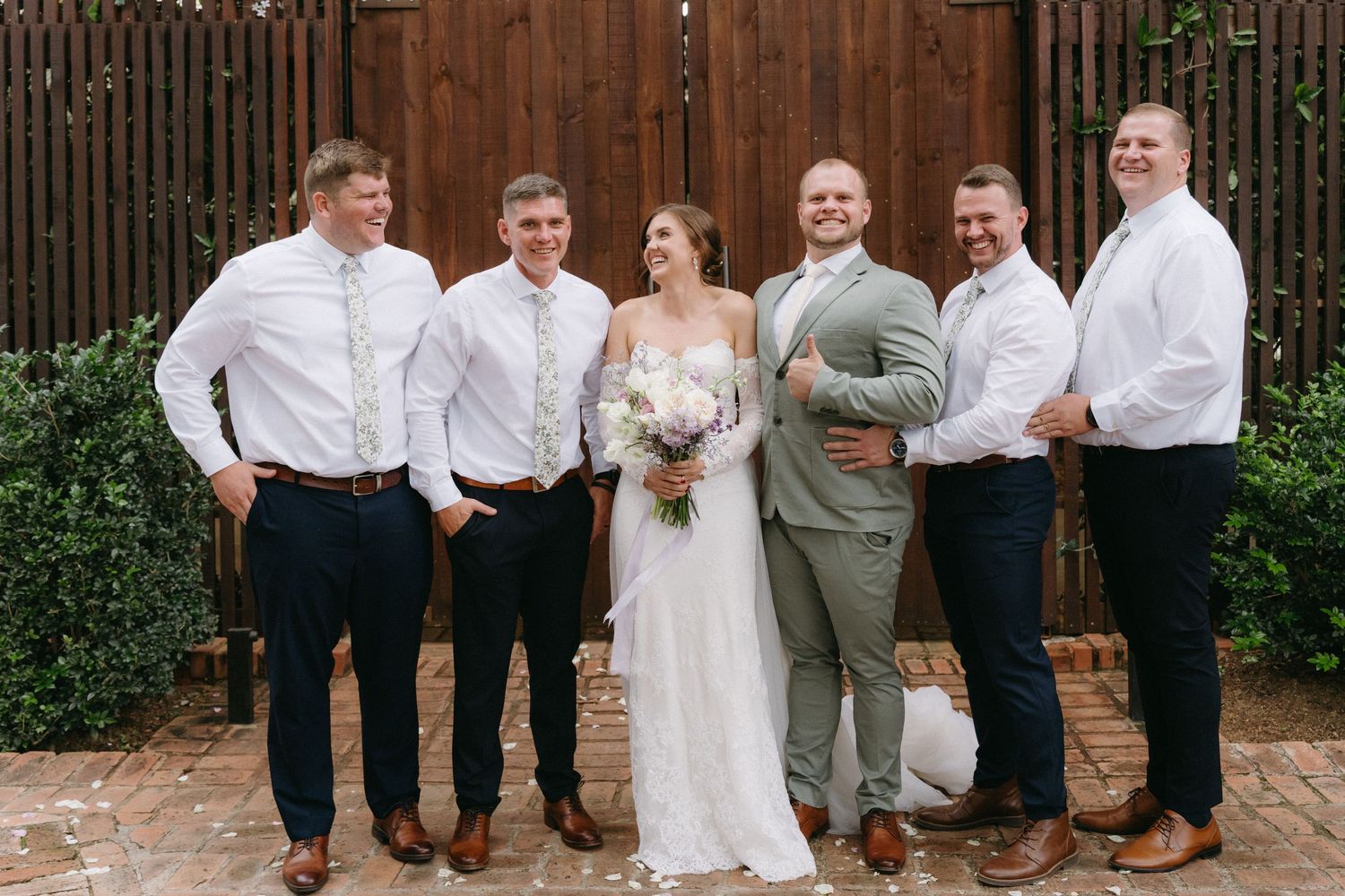 Wedding party in coordinated white and gray attire poses together against wooden fence backdrop.
