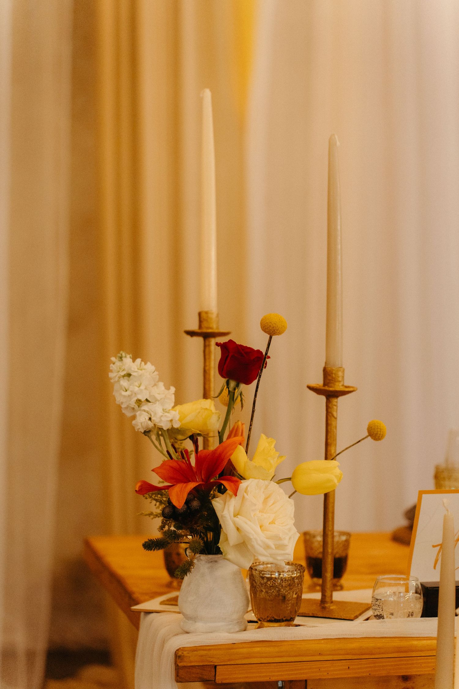 Elegant table setting with tall white candles and mixed floral arrangement in vases against a golden backdrop.
