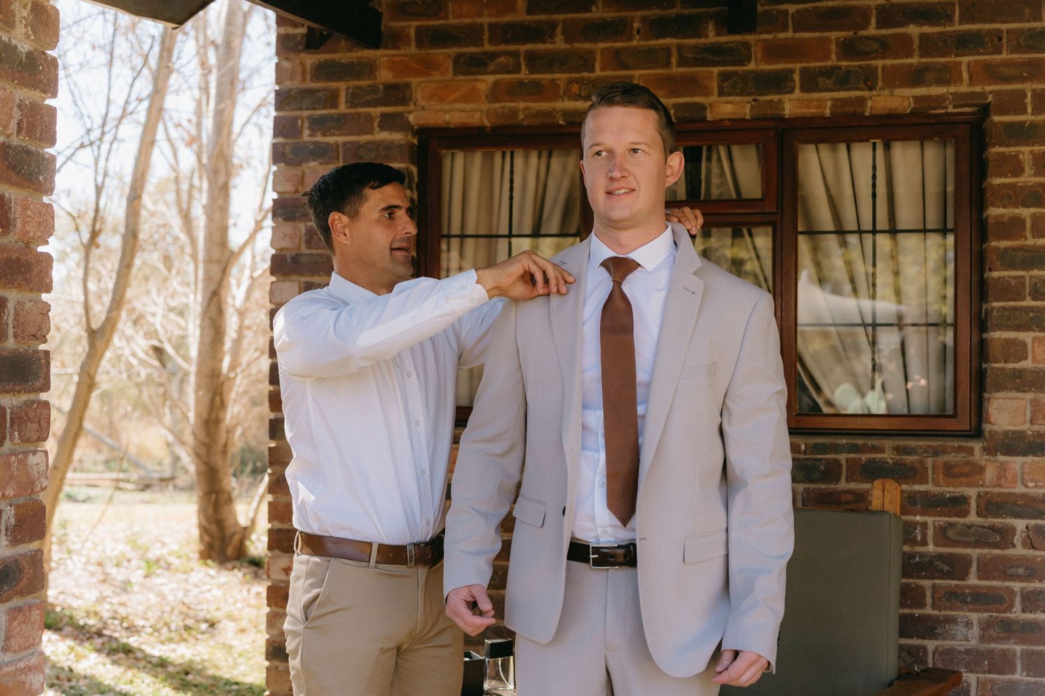 A groomsman helps adjust the groom's tie before the ceremony at a brick venue.