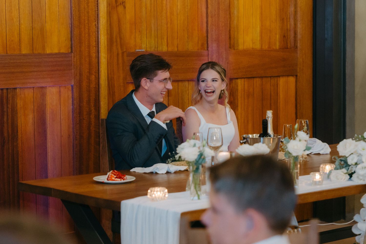 Newlyweds share an intimate moment while seated at their reception dinner table with wooden paneled walls.