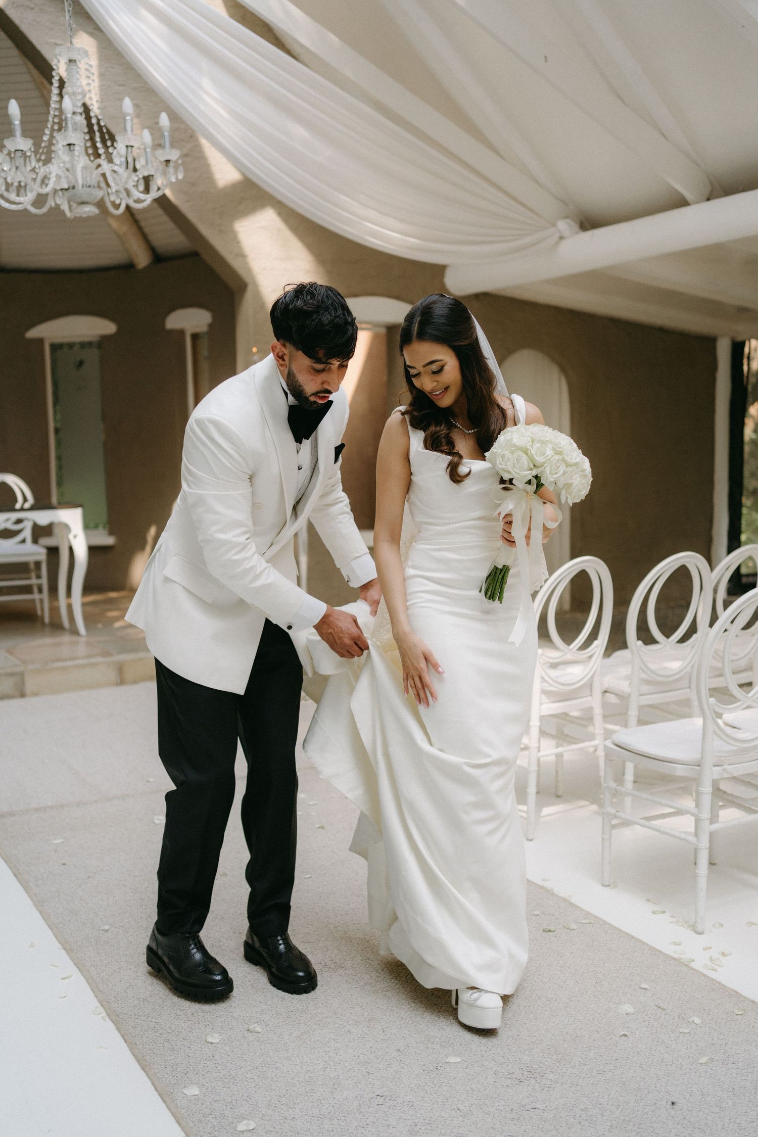 A couple in elegant wedding attire walking indoors with white draped ceiling and ghost chairs in the background.
