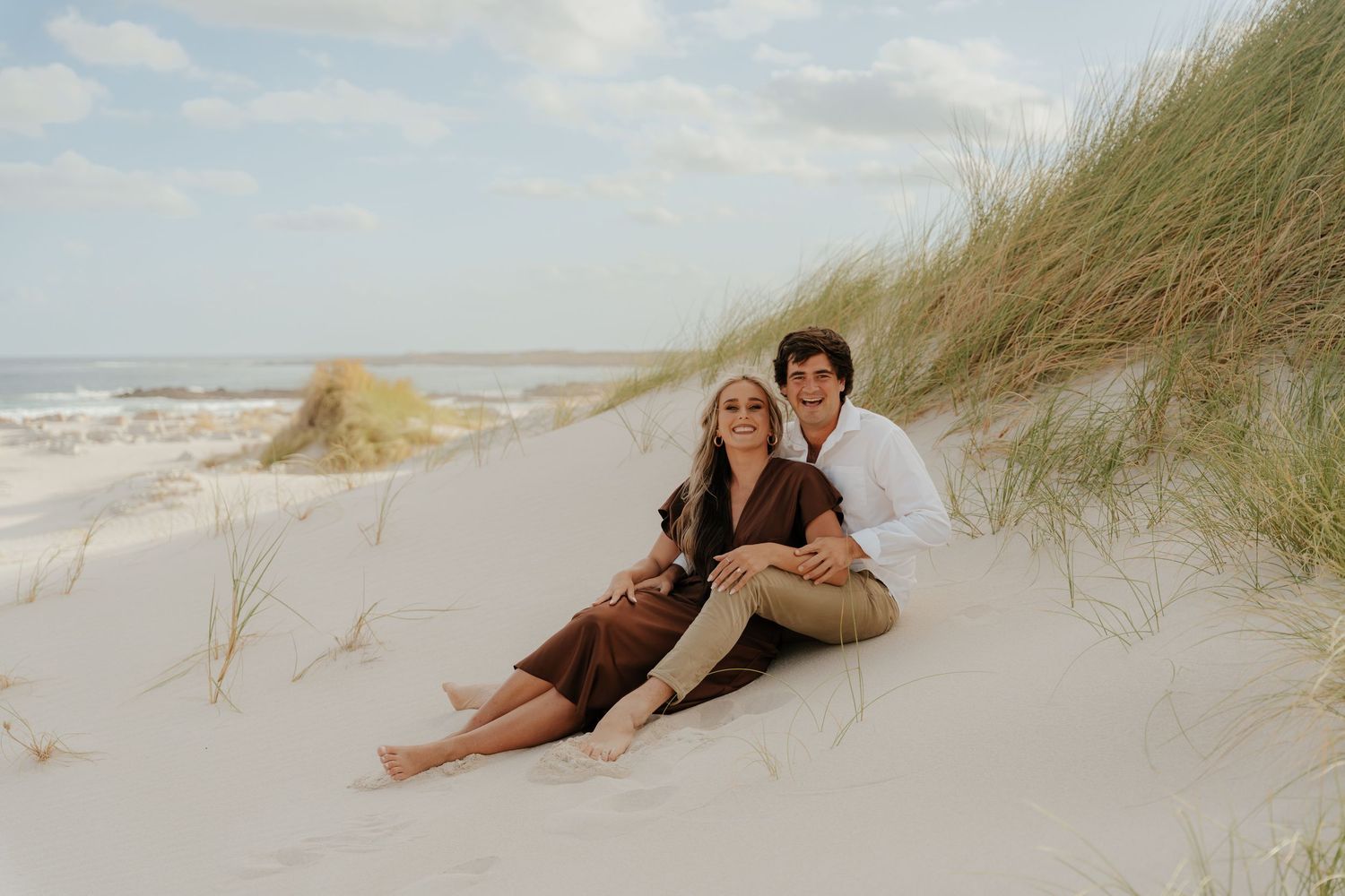Couple sitting on white sand dunes near the ocean, surrounded by beach grass on a sunny day.