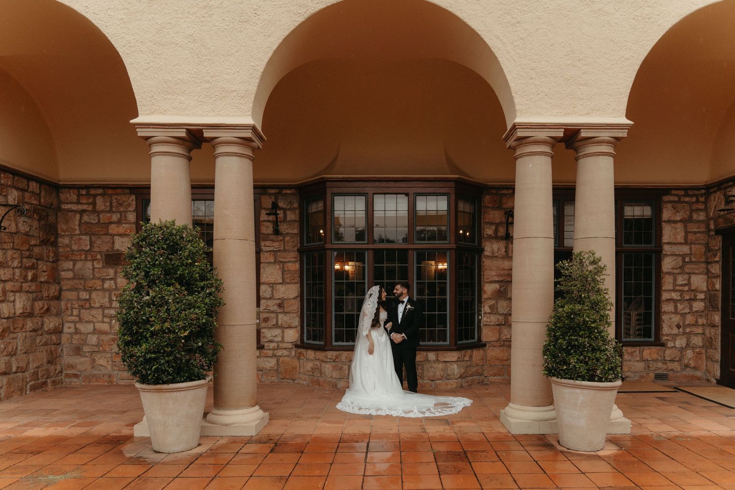 Bride and groom stand under arched colonnade of Glenshiel in Johannesburg on rainy wedding day.