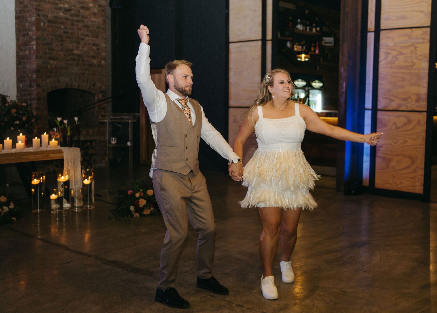 Couple dances energetically in reception venue with brick wall and candlelit fireplace in background.