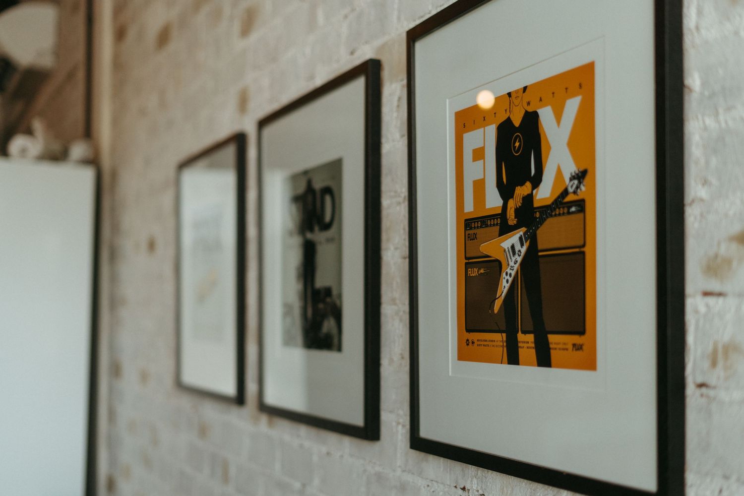 Three framed artworks hang on a textured white wall, with the rightmost frame containing an orange and black graphic design.
