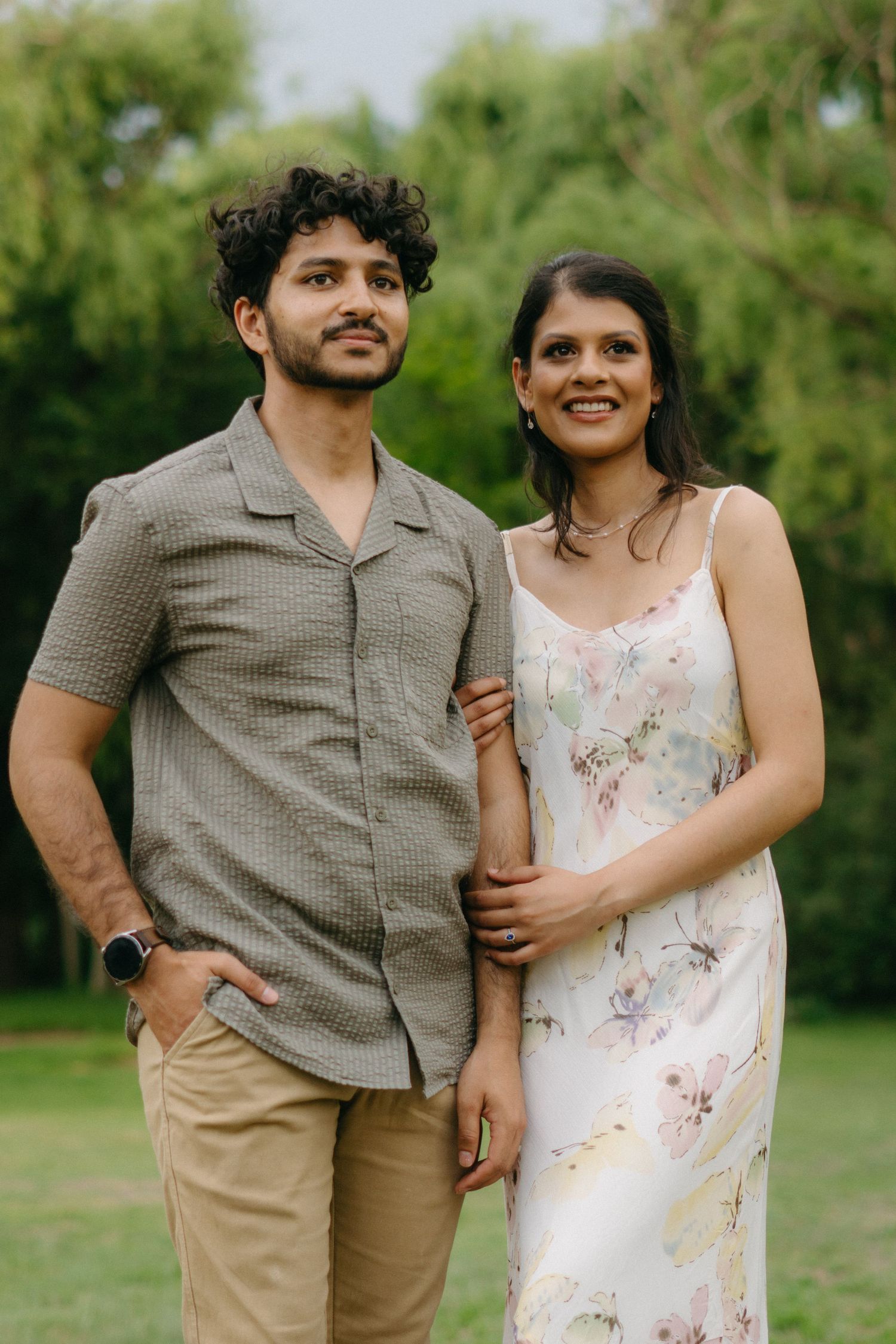 A loving couple poses outdoors in casual attire against a lush green background.