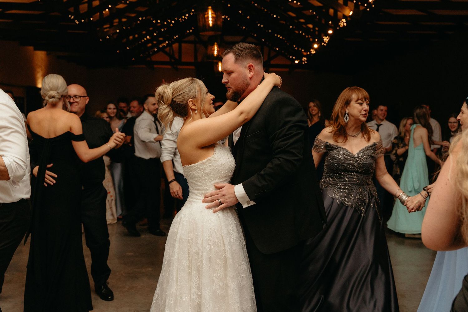 Wedding celebration with guests dancing and enjoying festivities in dimly lit rustic reception venue.
